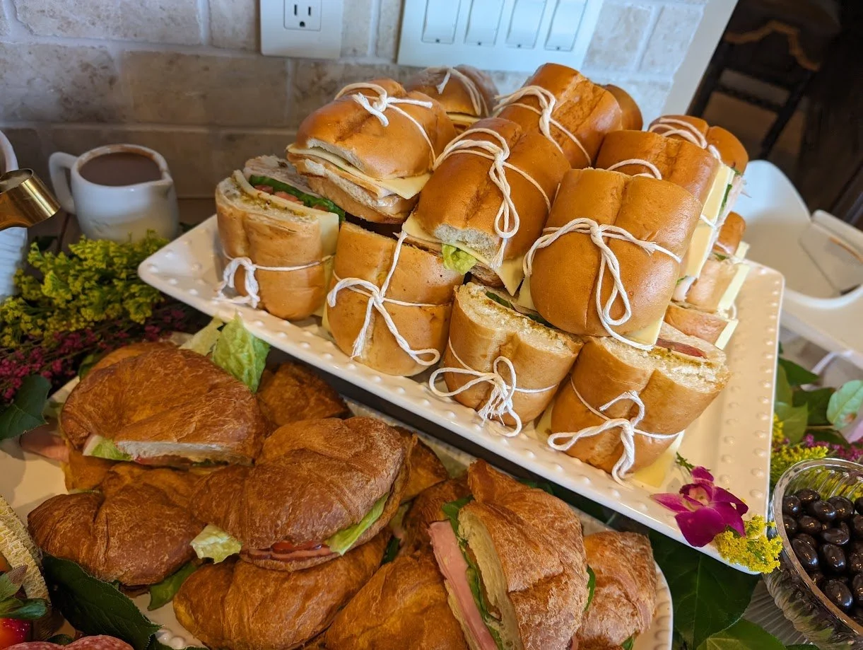 A platter of sandwich pinwheels tied with string, with croissant sandwiches and other assorted sandwiches on decorative trays, surrounded by flowers and berries.