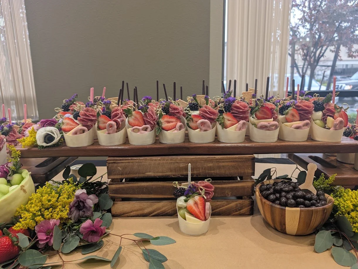 Dessert display with white cups filled with strawberries, blueberries, purple flowers, and edible decorations arranged on wooden trays, with a bowl of black olives and additional fruits and flowers on a table.