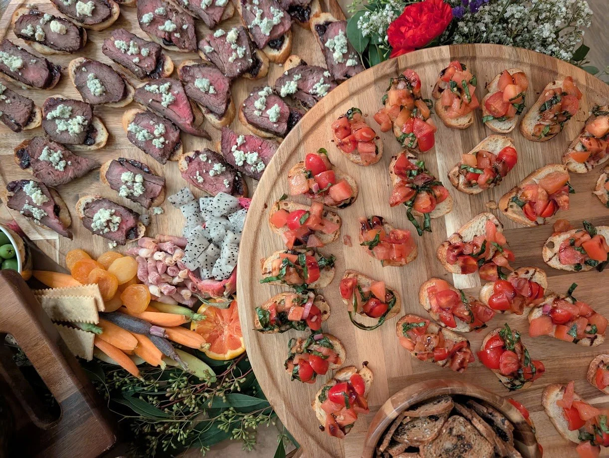 A wooden platter with various colorful bruschetta appetizers topped with tomatoes and herbs, surrounded by an assortment of sliced meats, cheeses, fruits, and vegetables, all arranged on a table with greenery and flowers in the background.