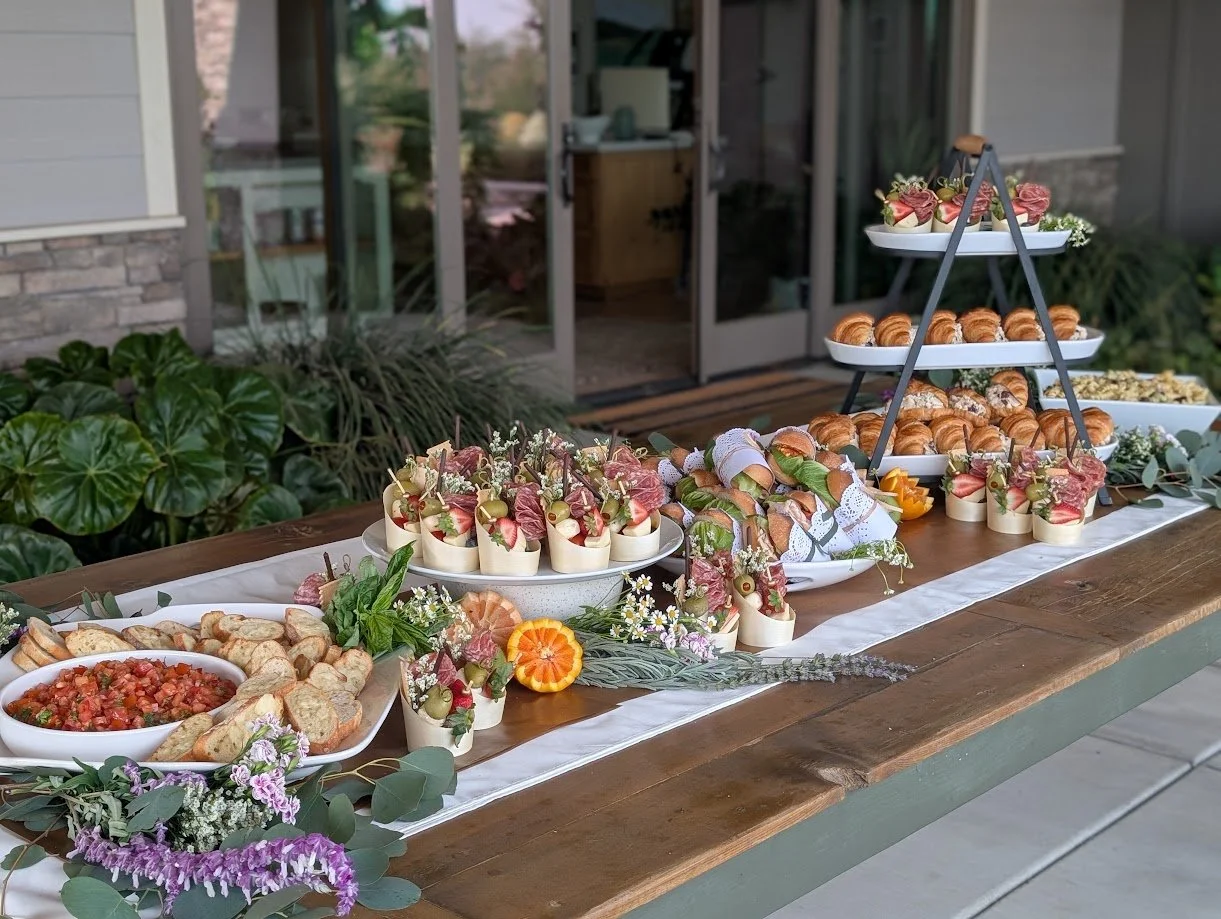 Outdoor table set with assorted appetizers, including sliced meats, cheeses, croissants, and dips, decorated with flowers and greenery.