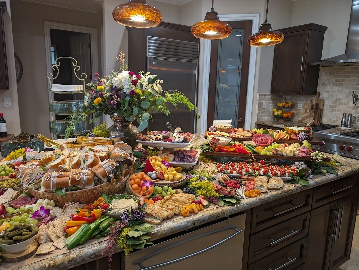A kitchen island laden with an assortment of food including bread, cheese, fruits, vegetables, and charcuterie, with a large floral arrangement in the center, illuminated by three copper pendant lights.