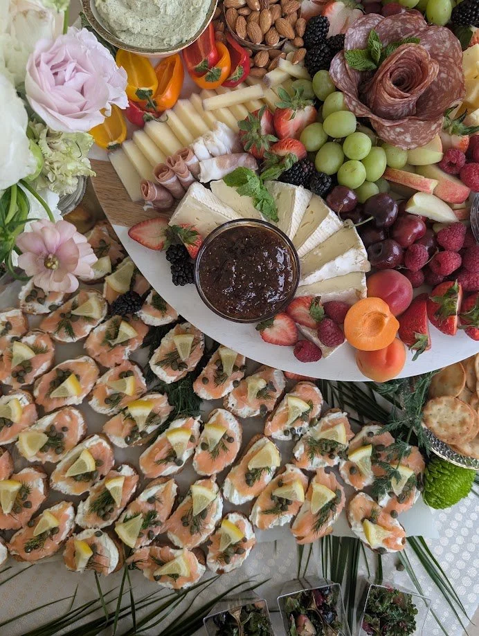 A platter of various cheeses, fresh fruits, nuts, and a tray of smoked salmon bites with lemon and herbs, part of an elaborate appetizer spread for a gathering.