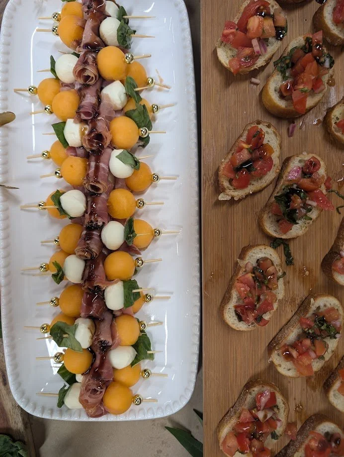 Assorted skewers with melon balls, mozzarella balls, prosciutto, and fresh basil on a white platter, and toasted bread slices topped with tomato bruschetta on a wooden board.