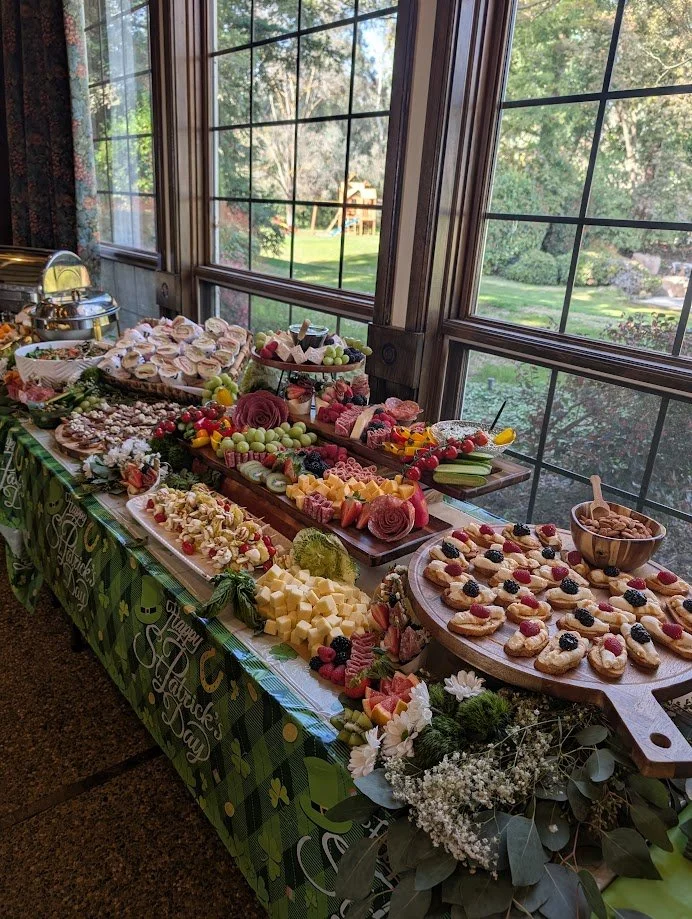 A table with a variety of desserts including fruit, candies, and cookies, set near large windows overlooking a green yard.