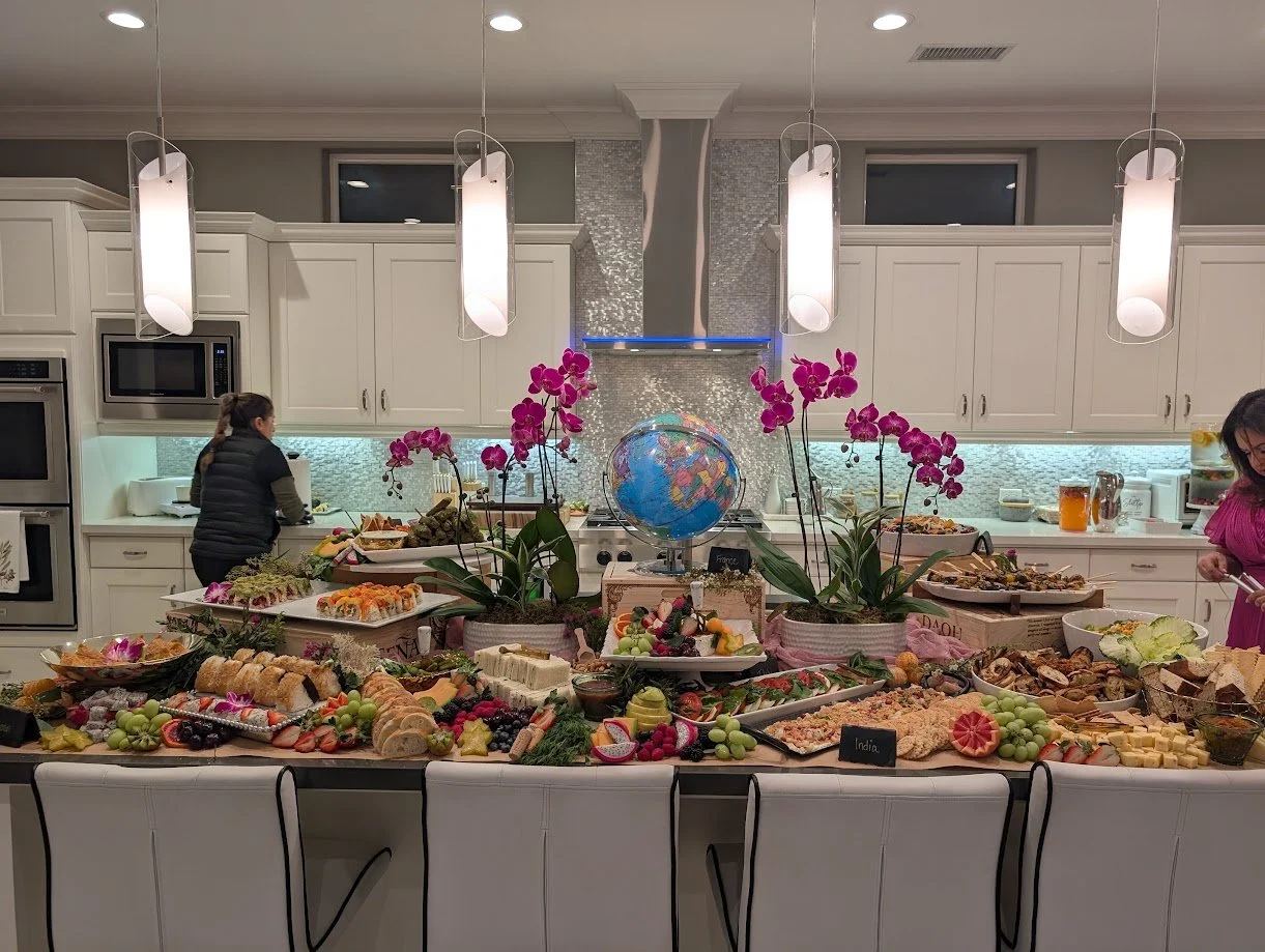 A buffet table with various international foods and an central globe, flanked by potted orchids, in a modern kitchen with white cabinets and pendant lighting.
