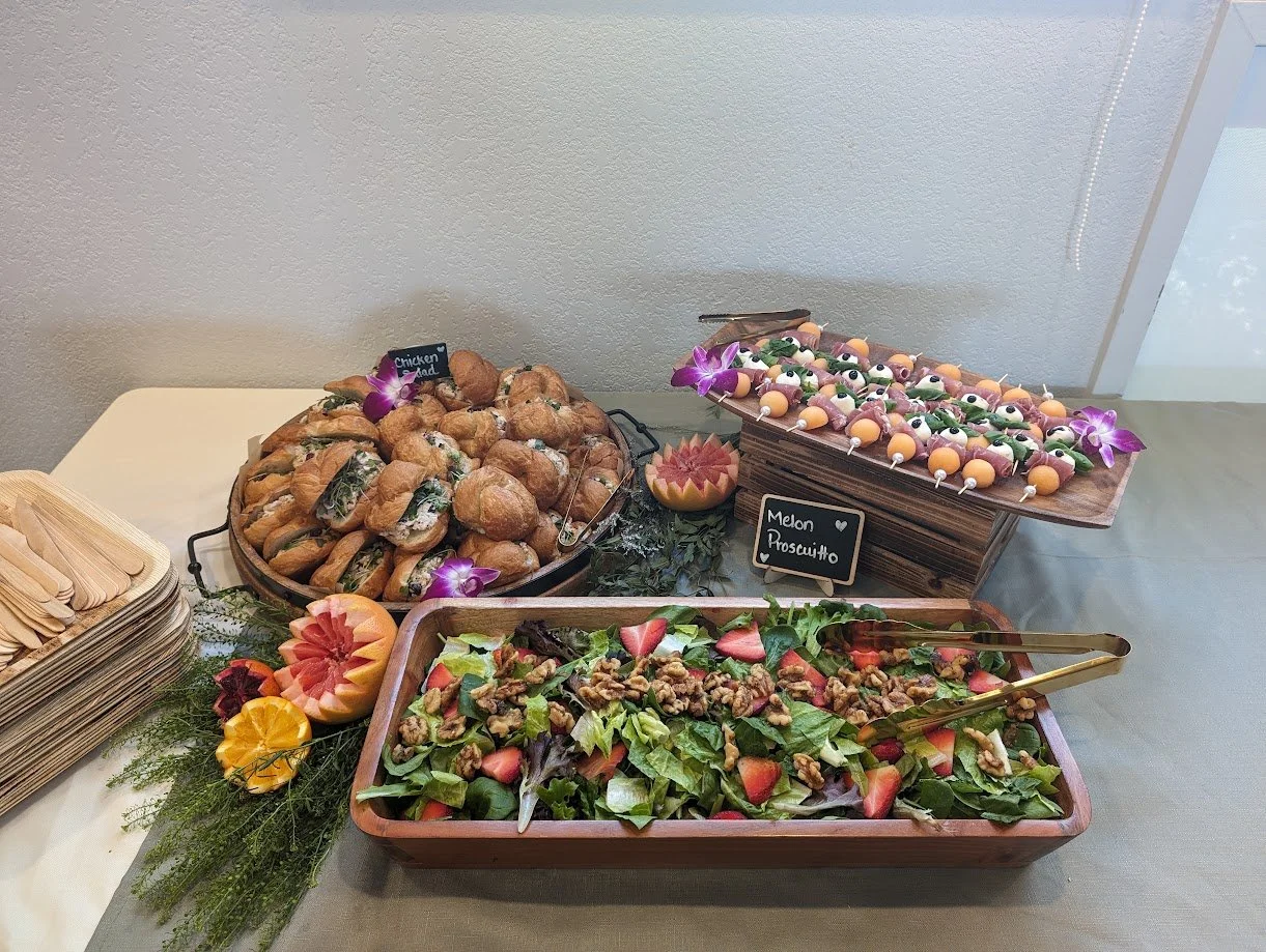 Buffet table with assorted finger foods including mini sandwiches, fruit skewers, a salad with strawberries and walnuts, and carved melons, decorated with purple orchids.