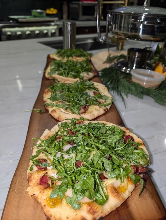 Three small flatbread pizzas topped with fresh arugula and bacon, served on a wooden board in a kitchen setting.