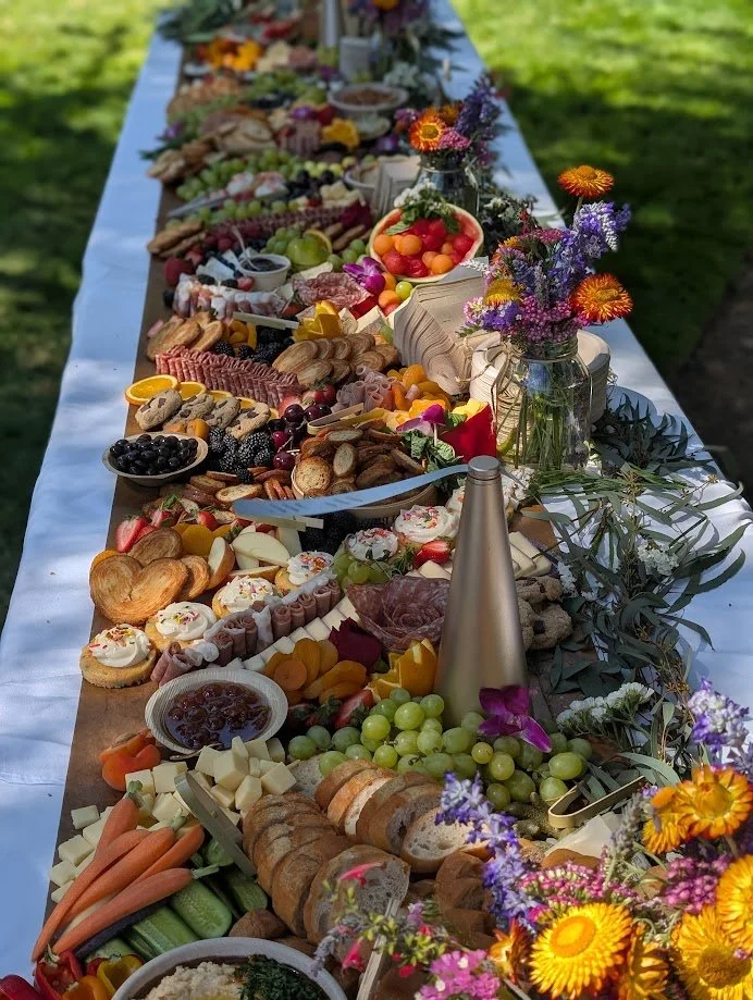 Long outdoor table filled with a variety of cheeses, fruits, vegetables, cookies, pastries, and floral arrangements for a celebration or gathering.