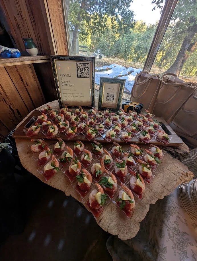 A tabletop with multiple pieces of appetizer in clear plastic cups, topped with slices of lemon, herbs, and what appears to be meat or fish. There are two framed signs on the table, one larger and one smaller, with QR codes and text mentioning 'Krystle & James Wedding.' The table is inside a rustic wooden room with large windows showing a landscape with trees and stamped snow outside.