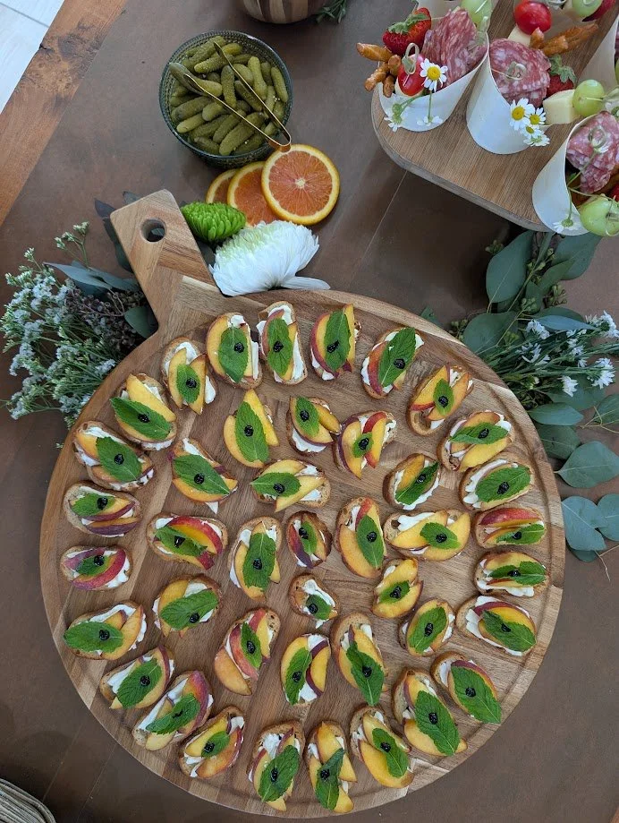 A large wooden platter with small canapé bites topped with fruit slices, mint leaves, and black olives, arranged for a gathering. There are also bowls of pickles, orange slices, and various snacks on the table.