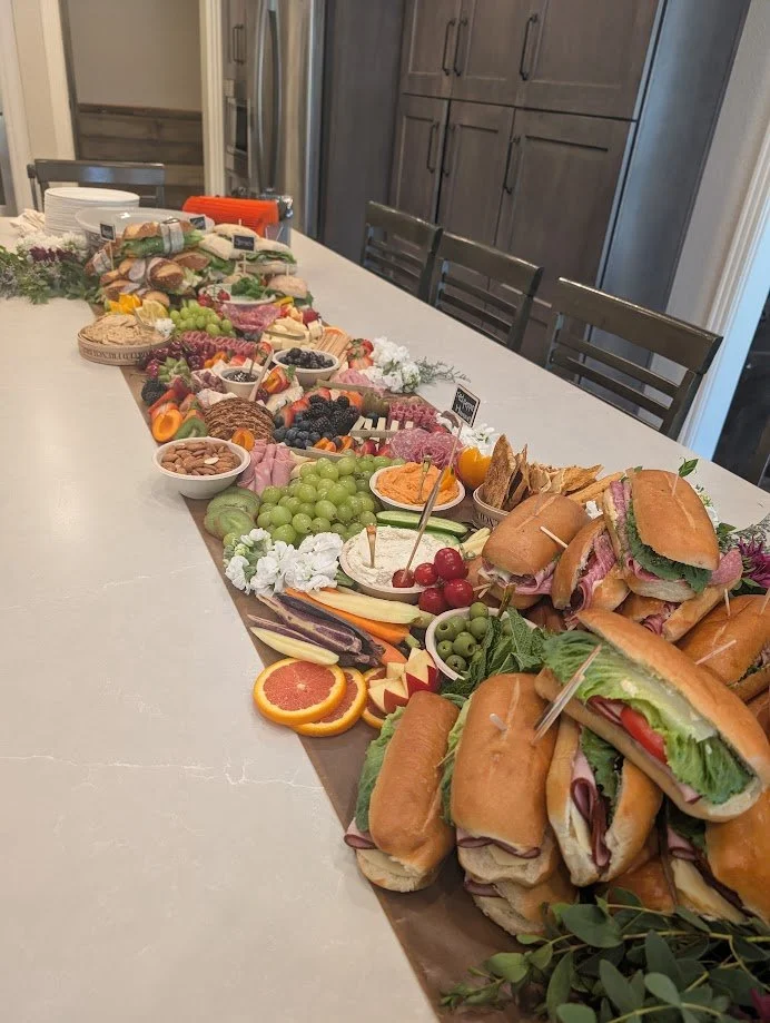 A long table set with a variety of sandwiches, fresh fruits, vegetables, cheese, and snacks for a party or gathering.