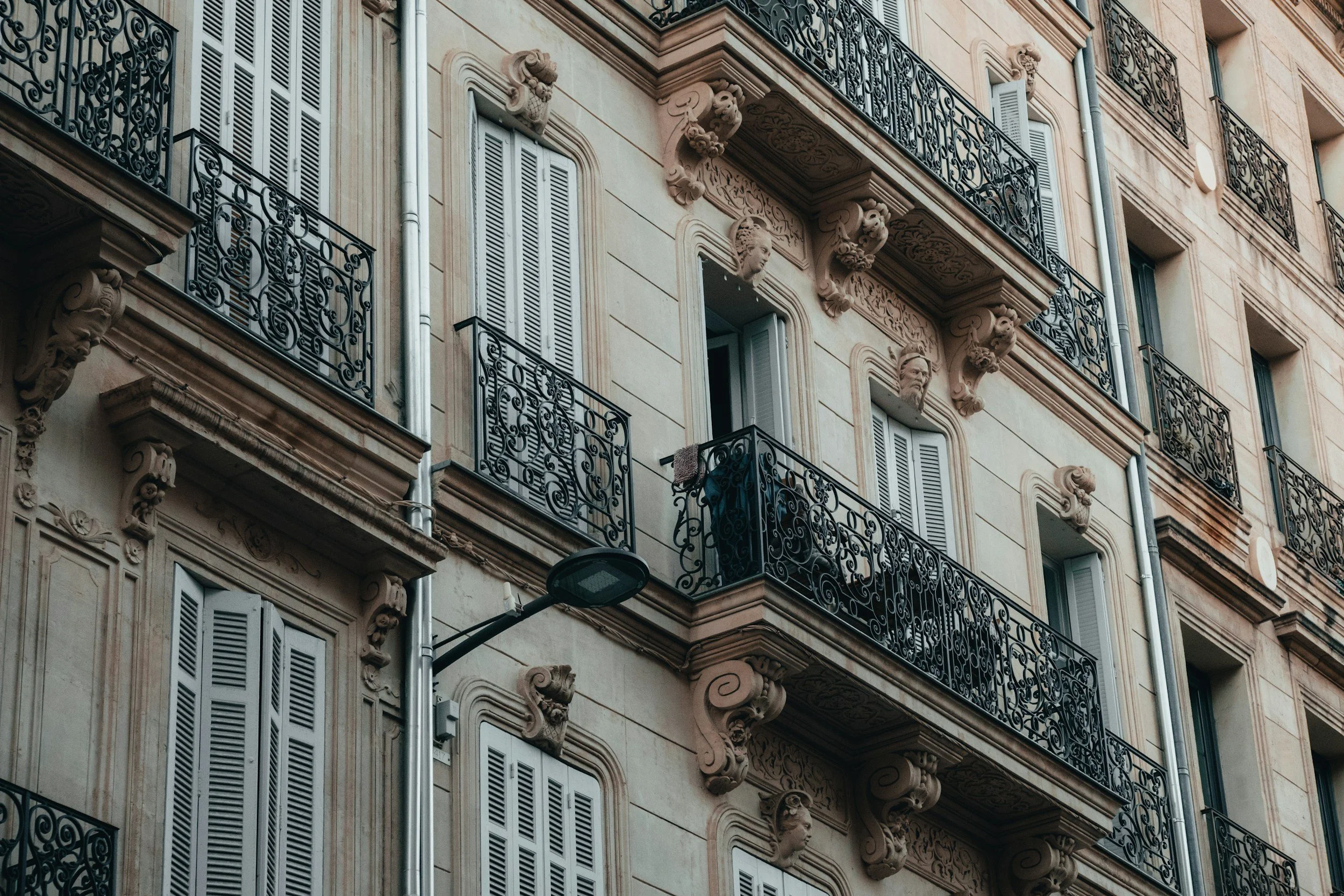 Close-up of a beige European style building facade with decorative stonework, ornate balcony railings, and closed shutters on the windows.