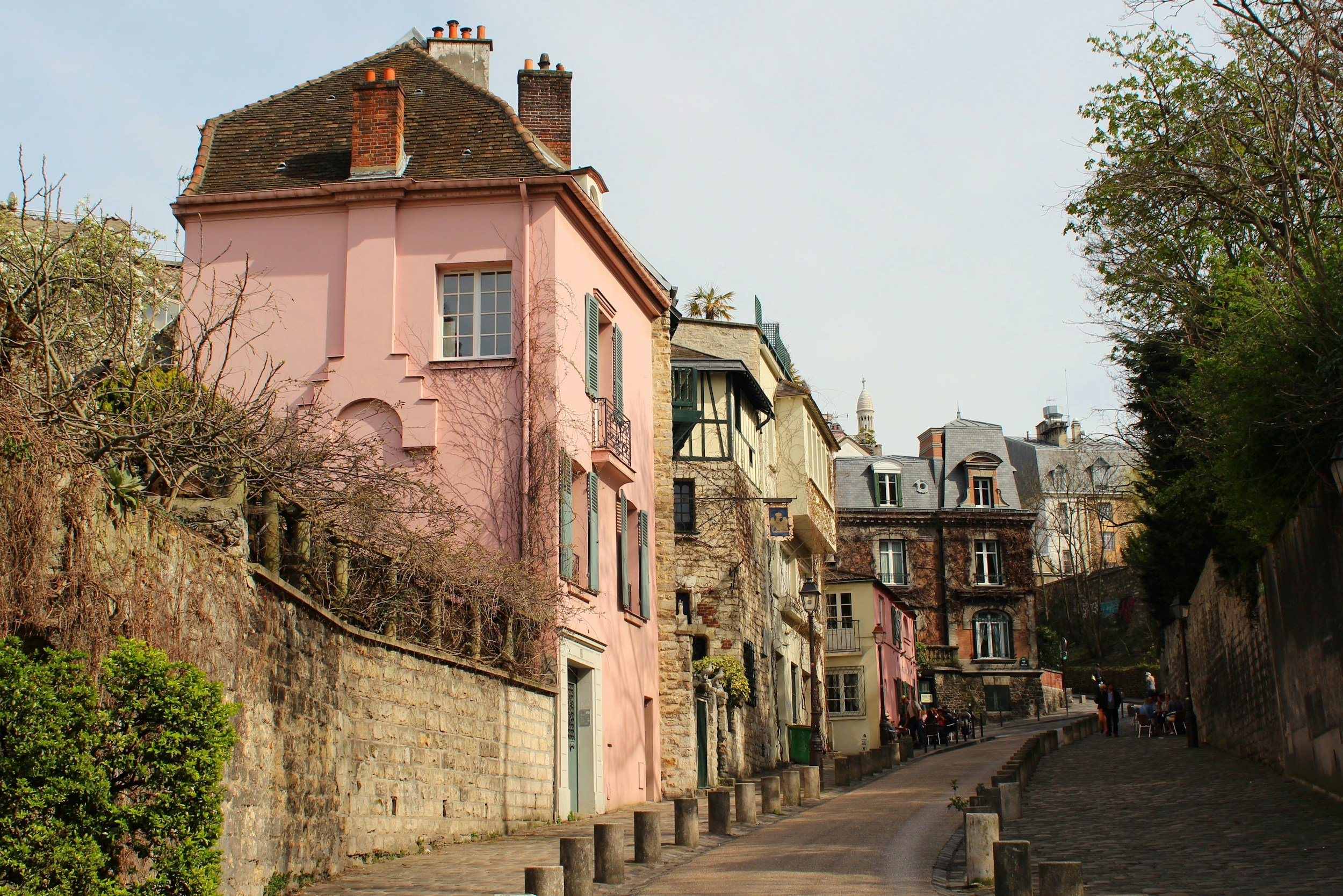 A French street lined with colorful houses, including a pink house with green shutters, a stone wall, trees, and a cloudy sky in the background.