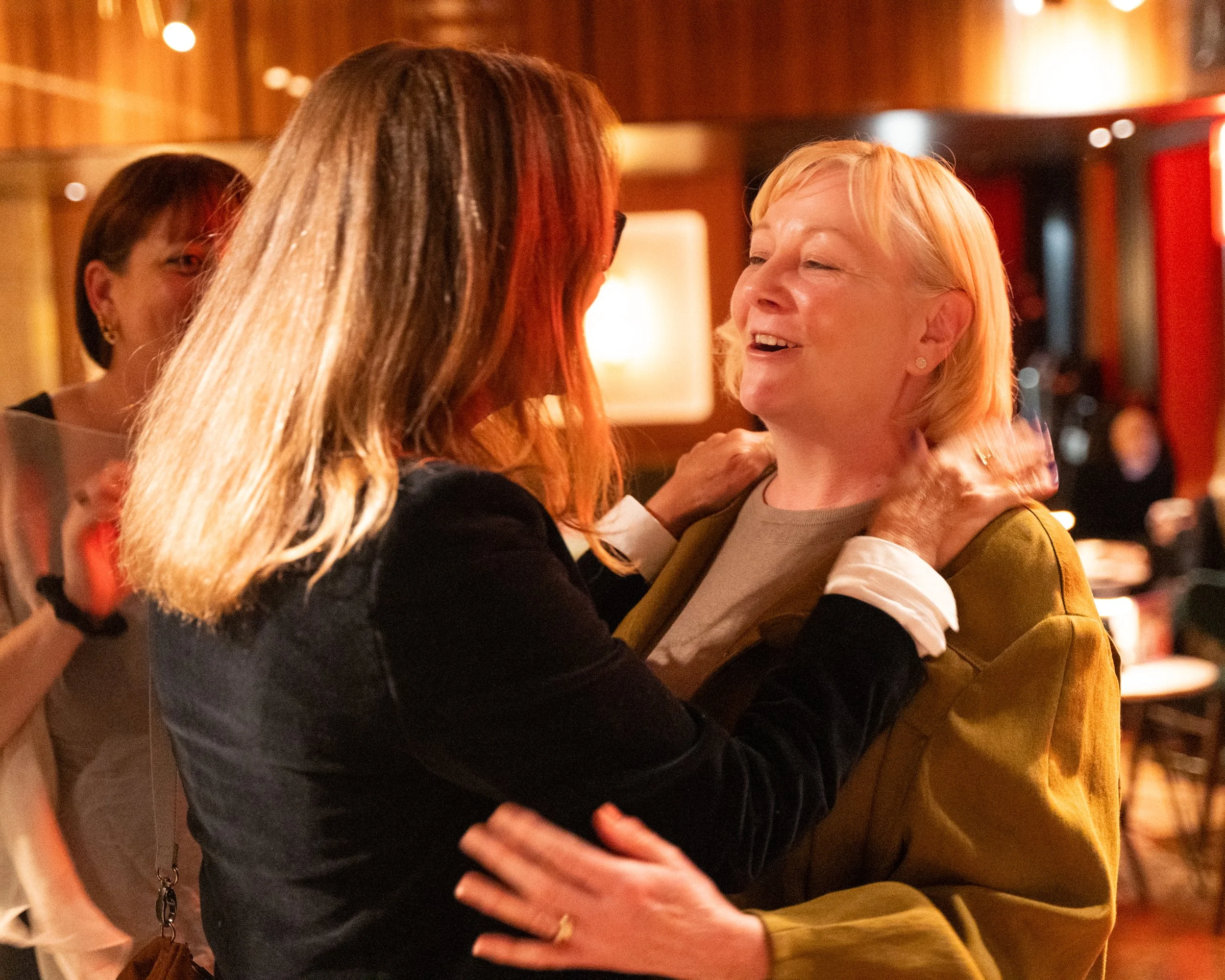 Two women are dancing closely, smiling, in a warmly lit indoor space.