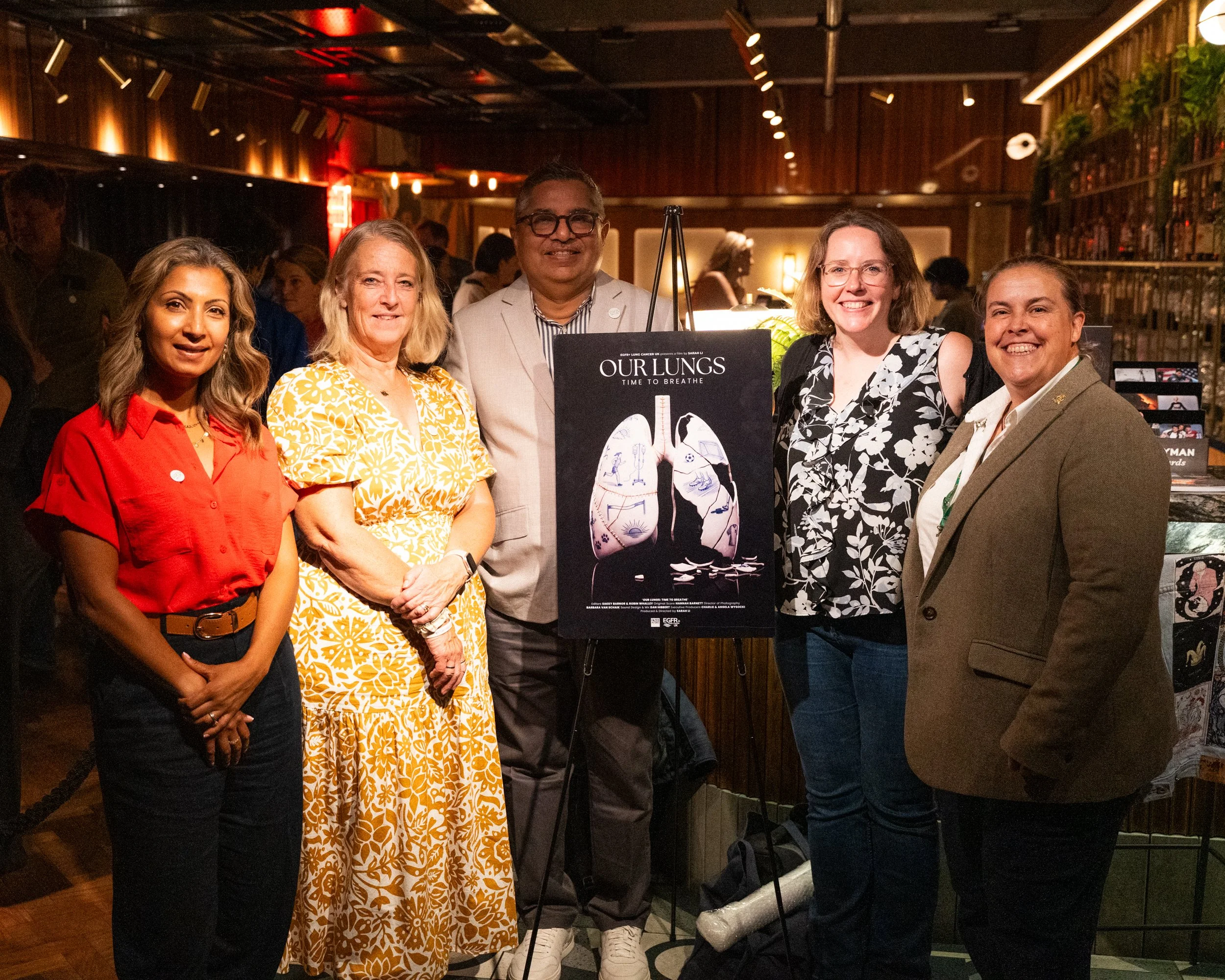 A diverse group of five people standing together indoors at a social event, smiling at the camera. They are around a poster with illustrations of lungs, with warm lighting and a wood-paneled background.