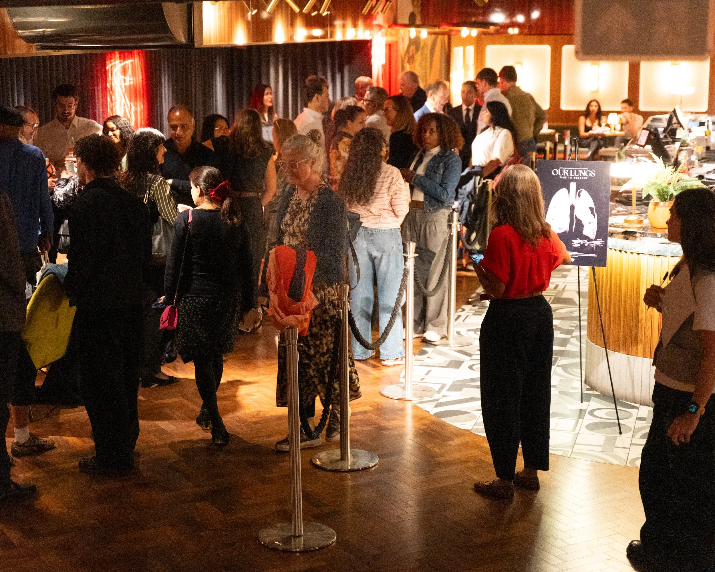 Crowd of people lined up at a restaurant or bar, waiting in line to order, with some chatting and others looking at menus or their phones. The setting is cozy and modern with warm lighting and a decorative background.