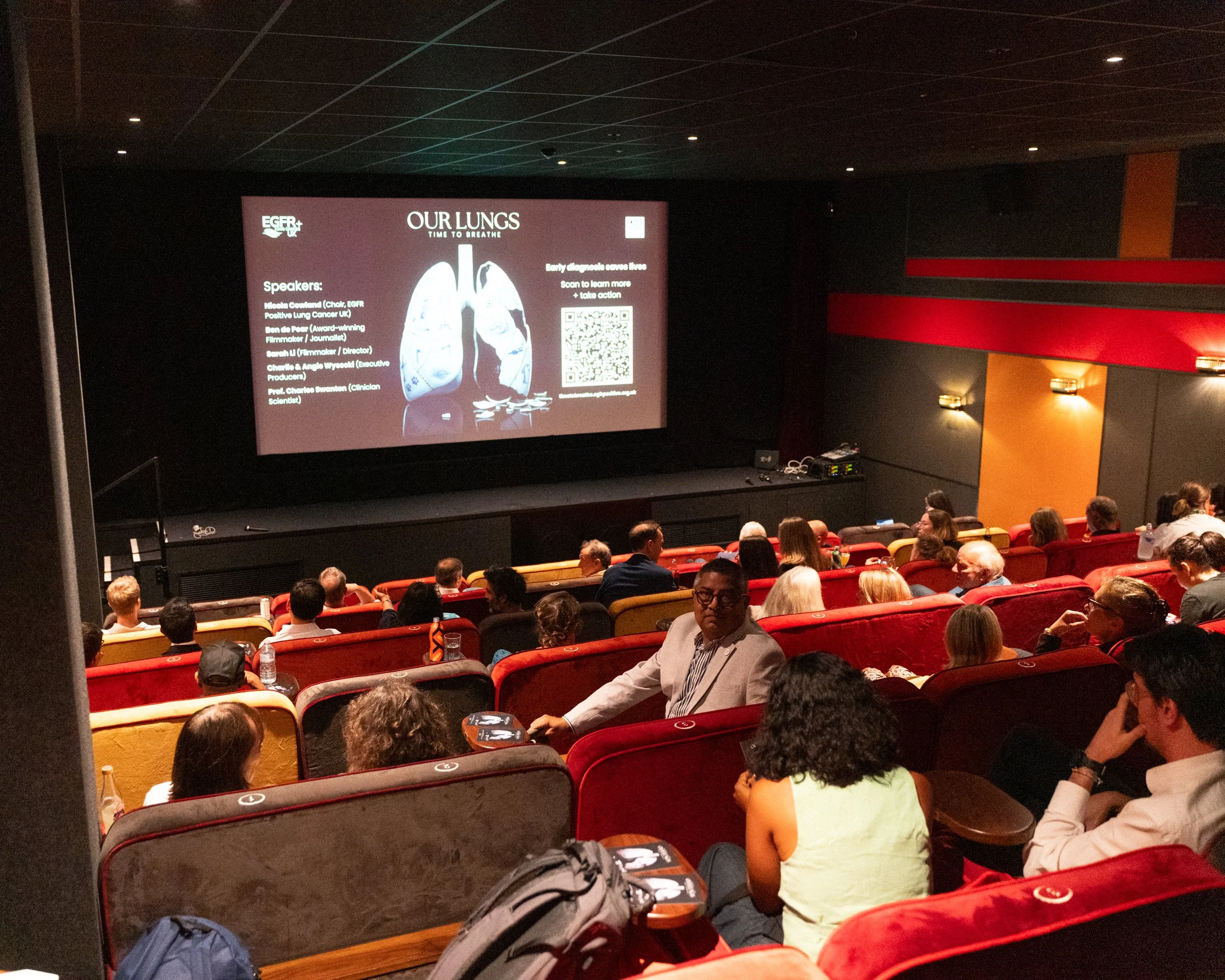 A darkened conference room filled with seated attendees watching a presentation on a large screen. The screen displays a slide titled "Our Lungs" with an illustration of lungs and text about a seminar related to respiratory health.