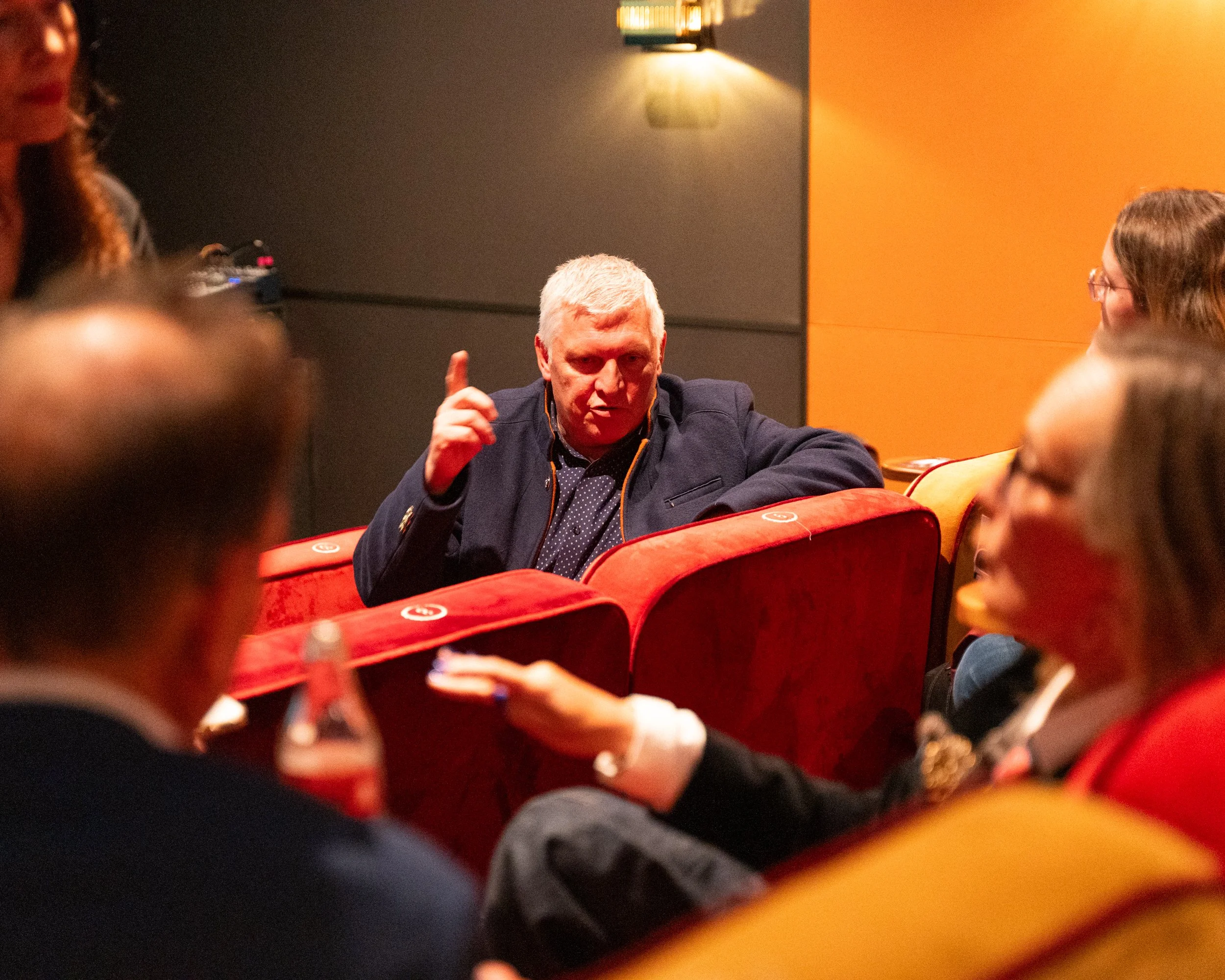A man with white hair gesturing with his index finger during a discussion at a meeting or conference, with attendees blurred in the foreground and colorful walls in the background.
