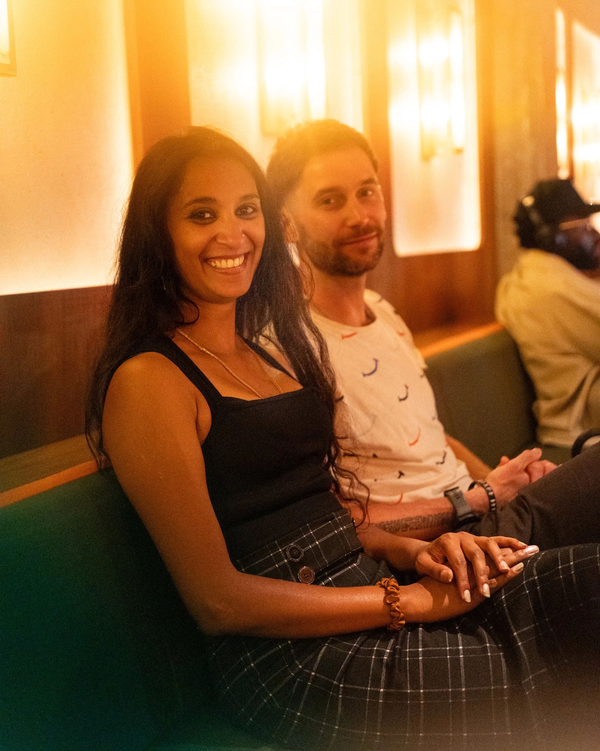 Two smiling people, a woman with long black hair and a man with short dark hair, sitting at a table in a warmly-lit room, possibly a restaurant or cafe.