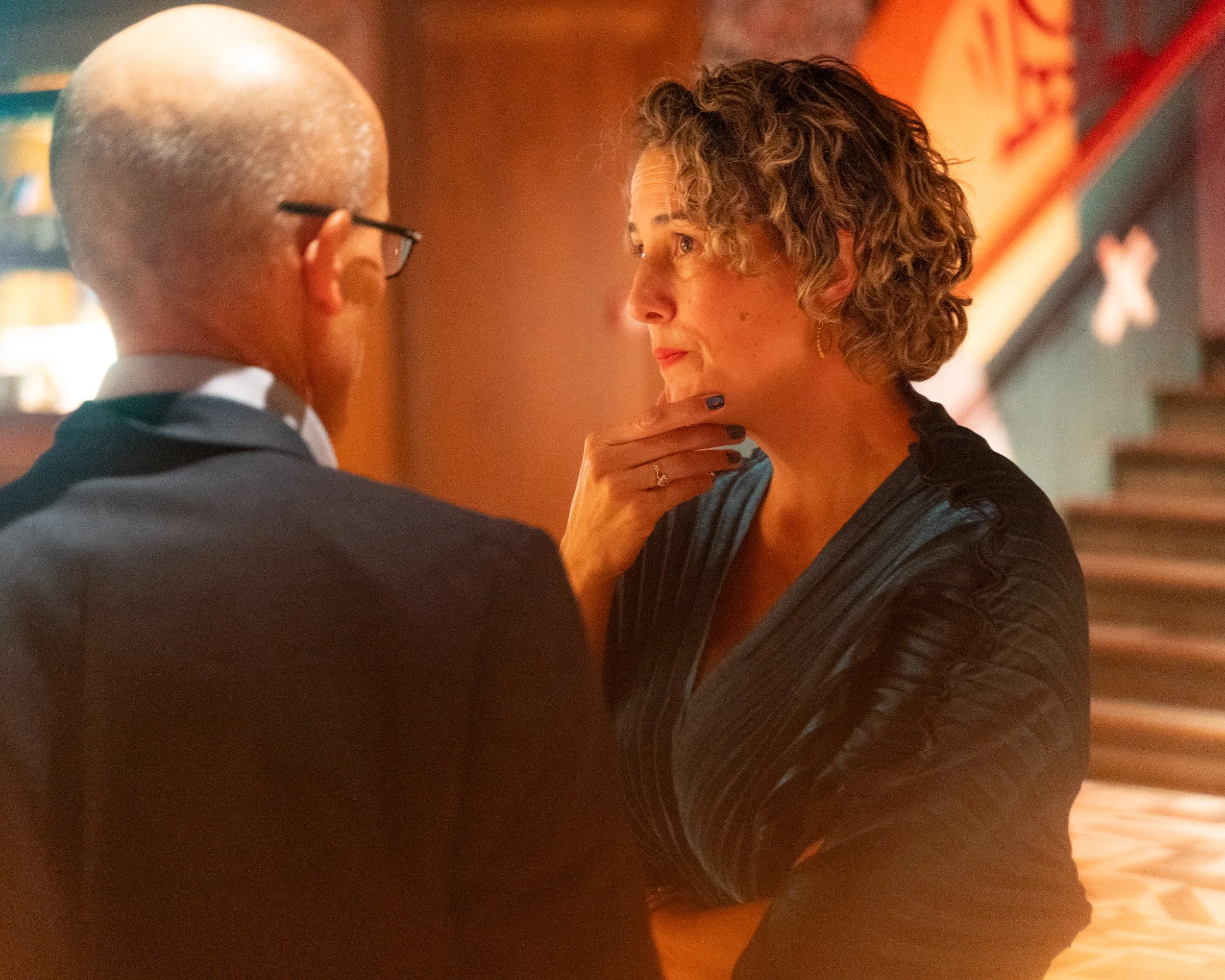 A woman with curly hair and hoop earrings touching her chin while talking to a man with glasses and bald head in a warmly lit room.