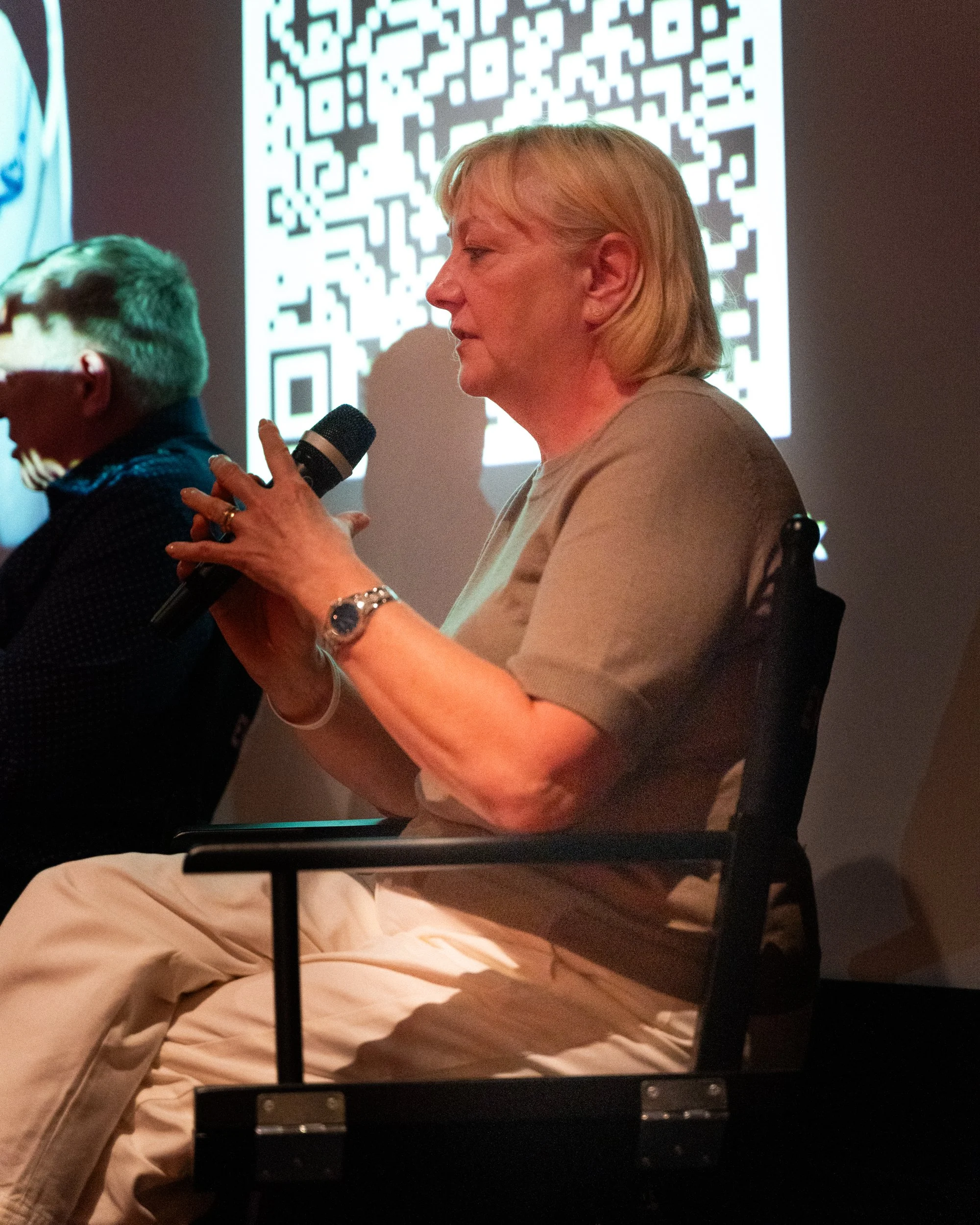 A woman with blonde hair and a beige shirt is sitting and speaking into a microphone, with a large QR code projected on the wall behind her. She is wearing a watch and a bracelet on her left wrist, and her eyes are closed.