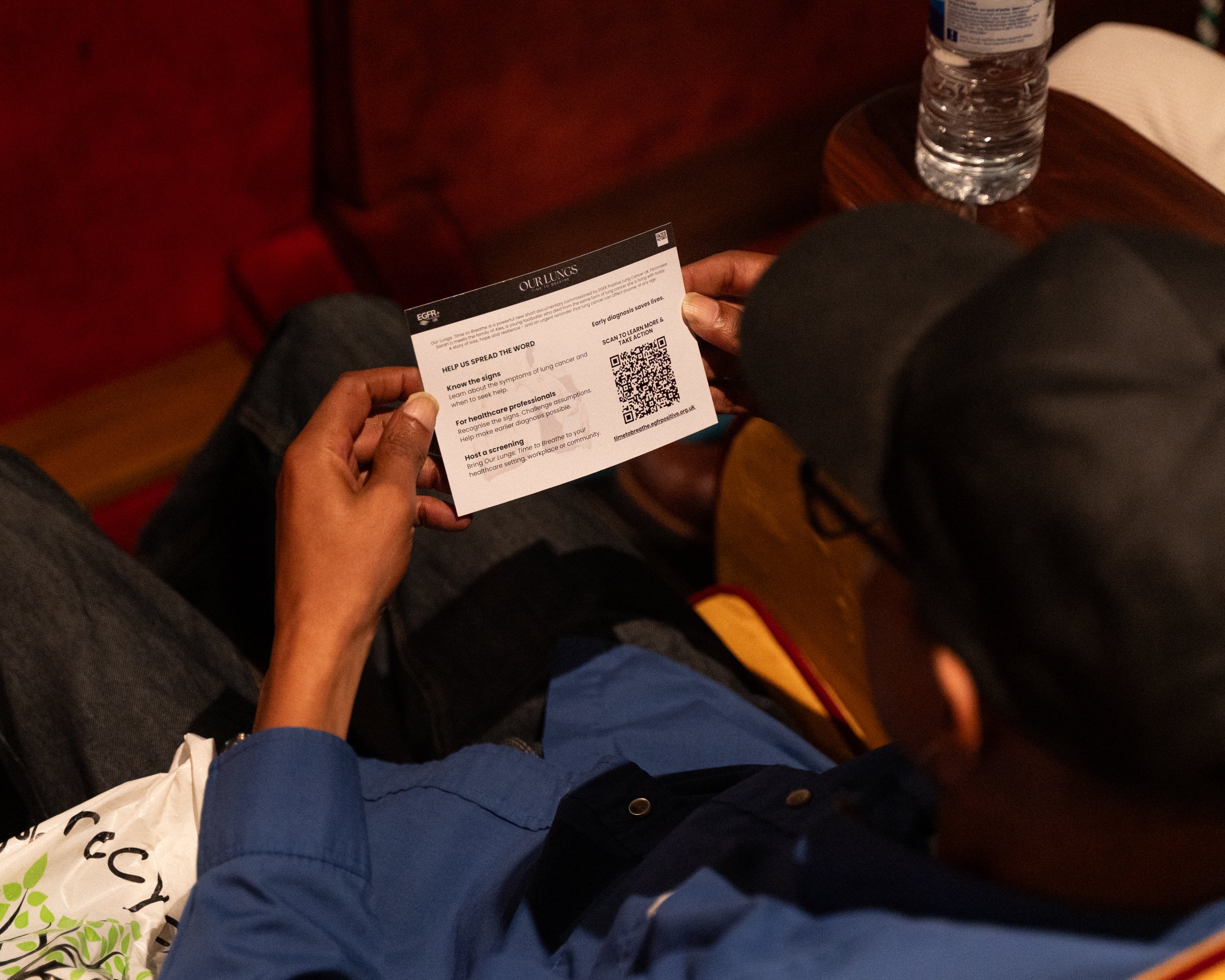 Person holding a printed card with text and a QR code, sitting at a wooden table with a water bottle and a paper napkin.