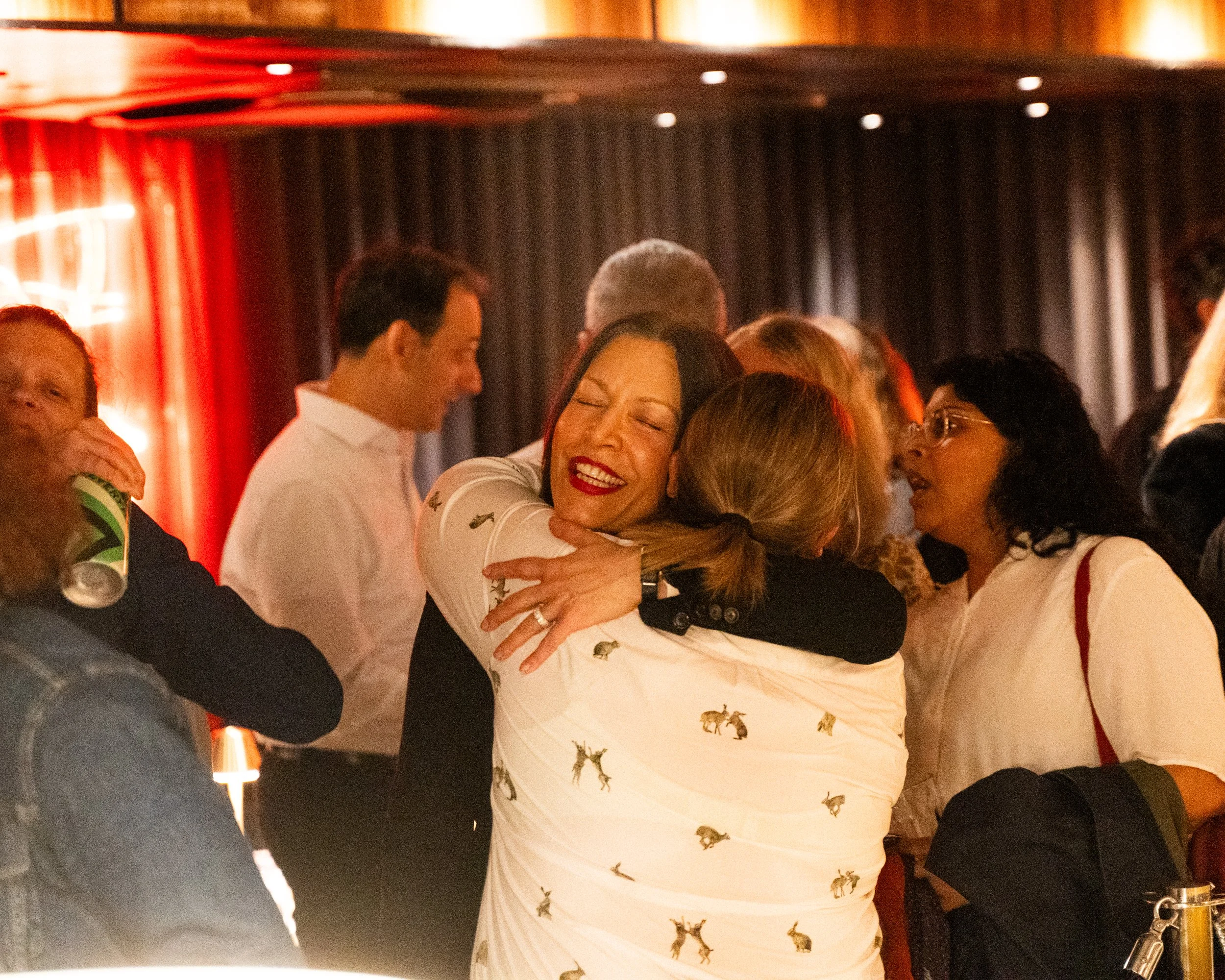 People celebrating, hugging, and enjoying a social event in a dimly lit venue with dark curtains.