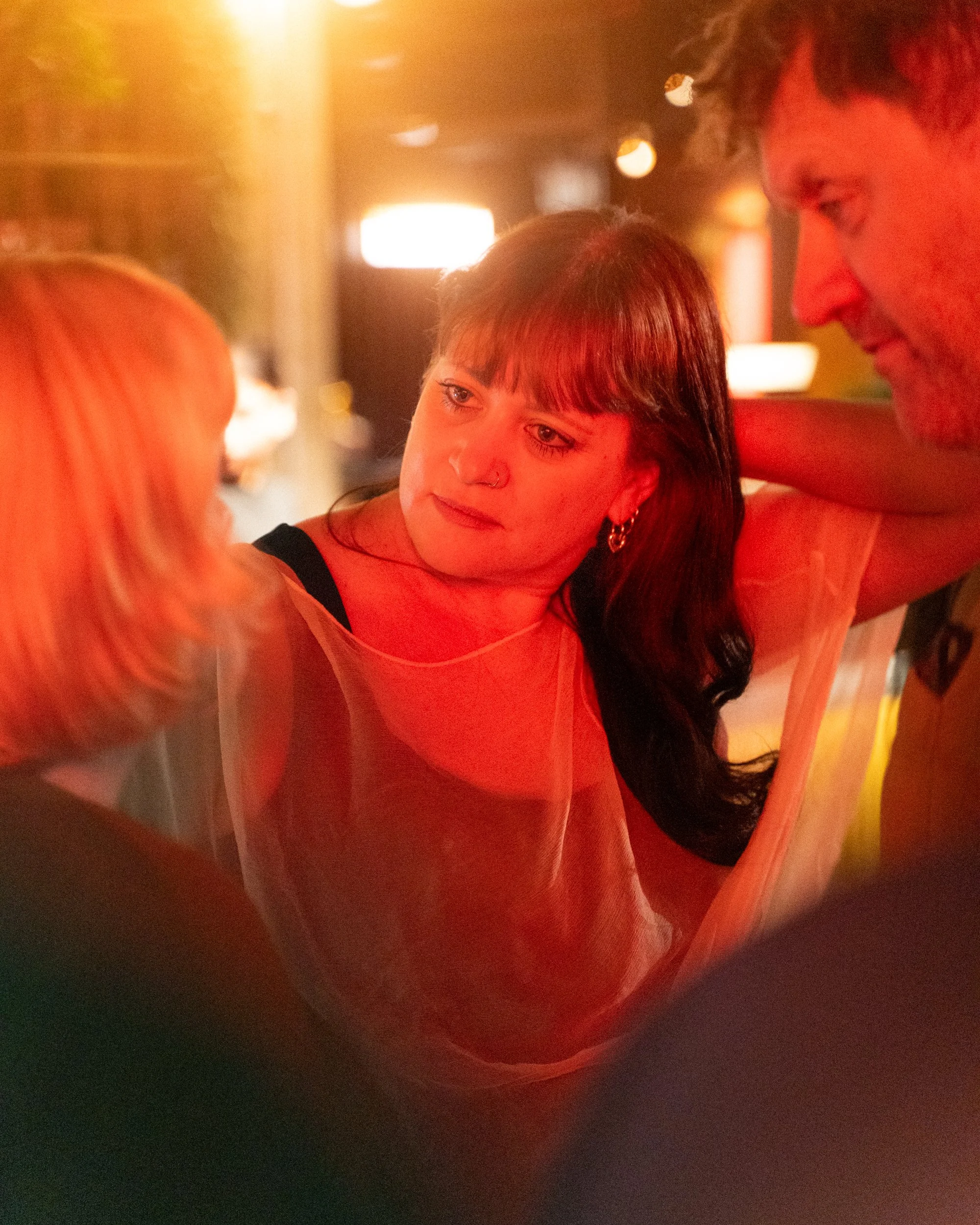 A young woman and man are looking at each other closely, smiling, inside a warmly lit room with blurry lights in the background.
