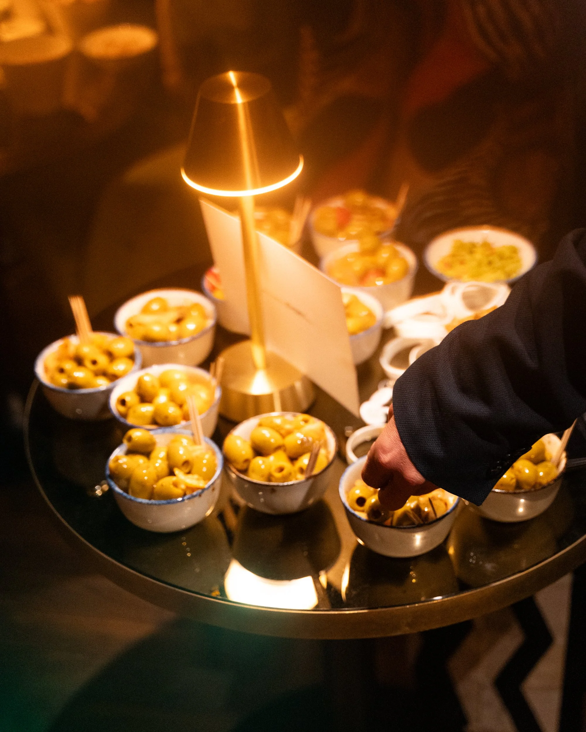 A person placing green olives with sticks into small white bowls arranged around a central heat source, likely at a buffet or food station.