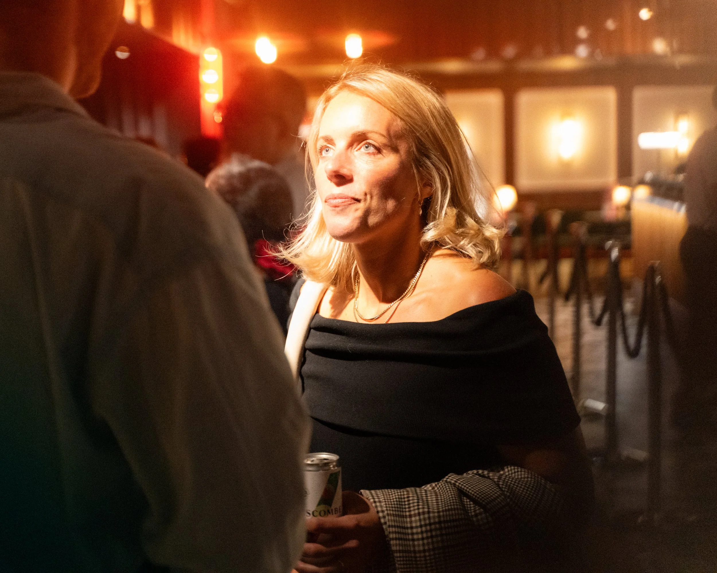 Woman with blonde hair and a black top, looking upwards in a dimly lit indoor setting with warm lighting and blurred background.