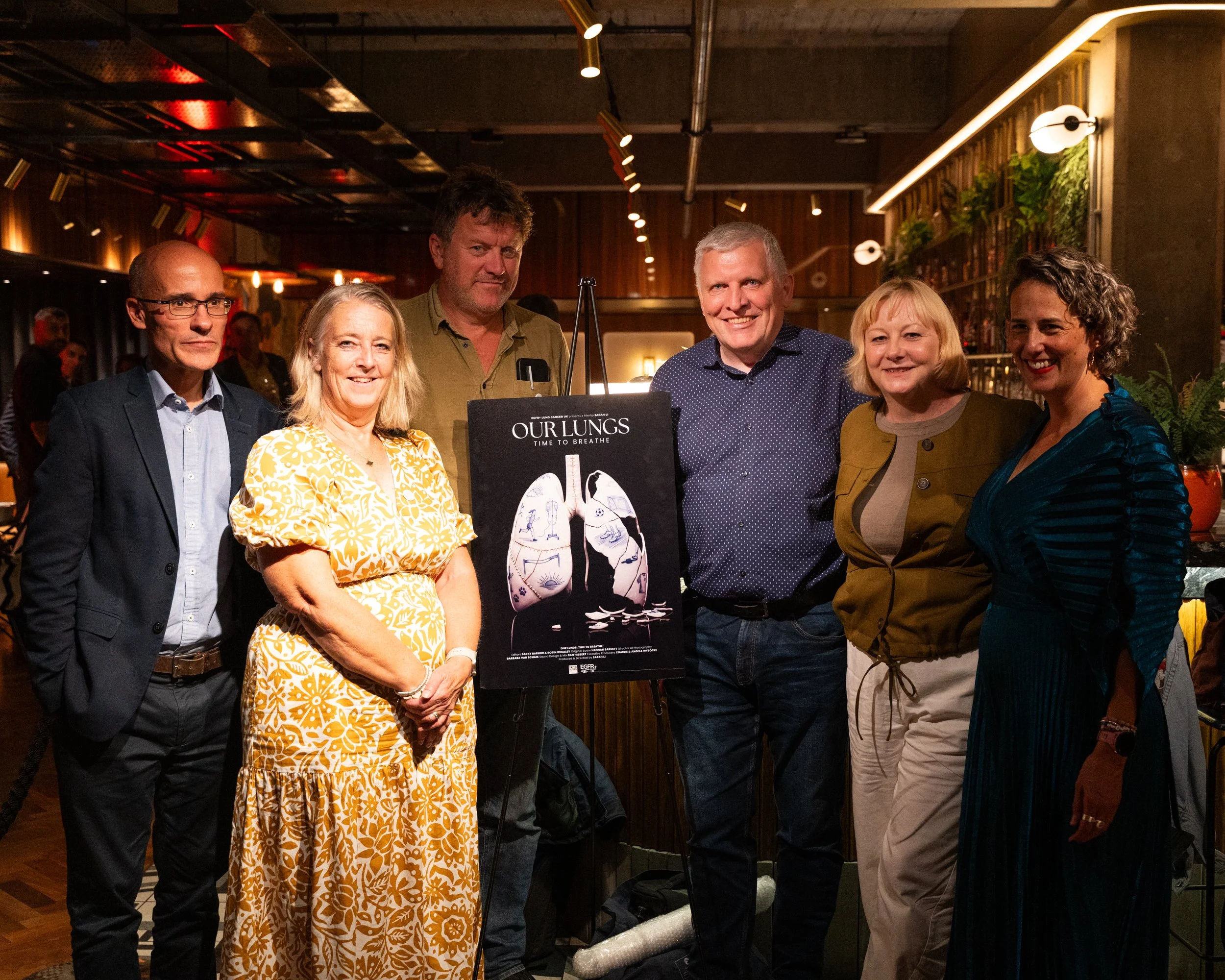 Group of seven people standing together at an indoor event, smiling for the photo. There are three women and four men, and a sign promoting lung health is visible in the middle.