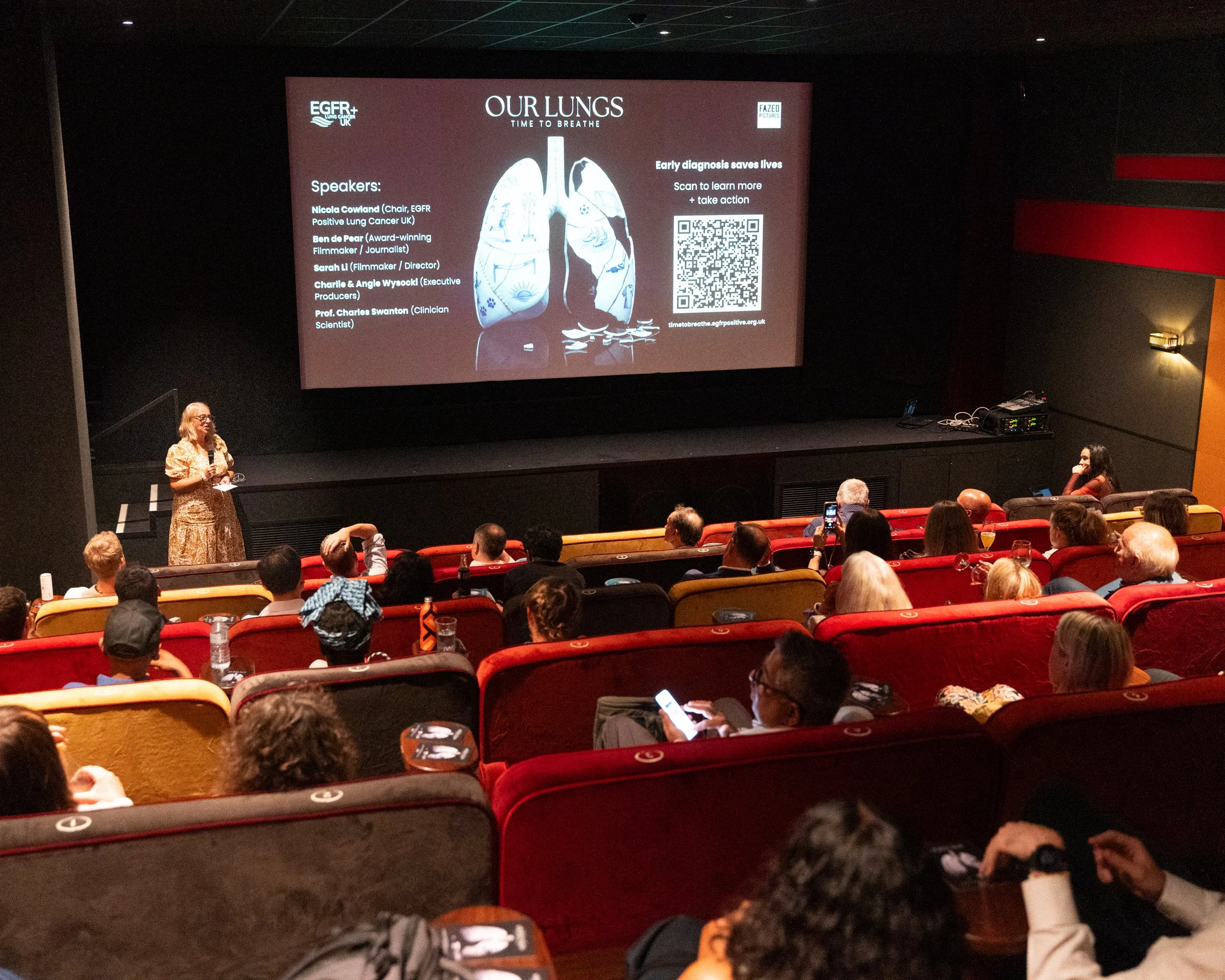 A woman presenting on stage at a conference or event, with a large screen behind her that displays a slide titled "OUR LUNGS: TIME TO BREATHE" and information about lung health and early diagnosis. The audience is seated in theater-style chairs.
