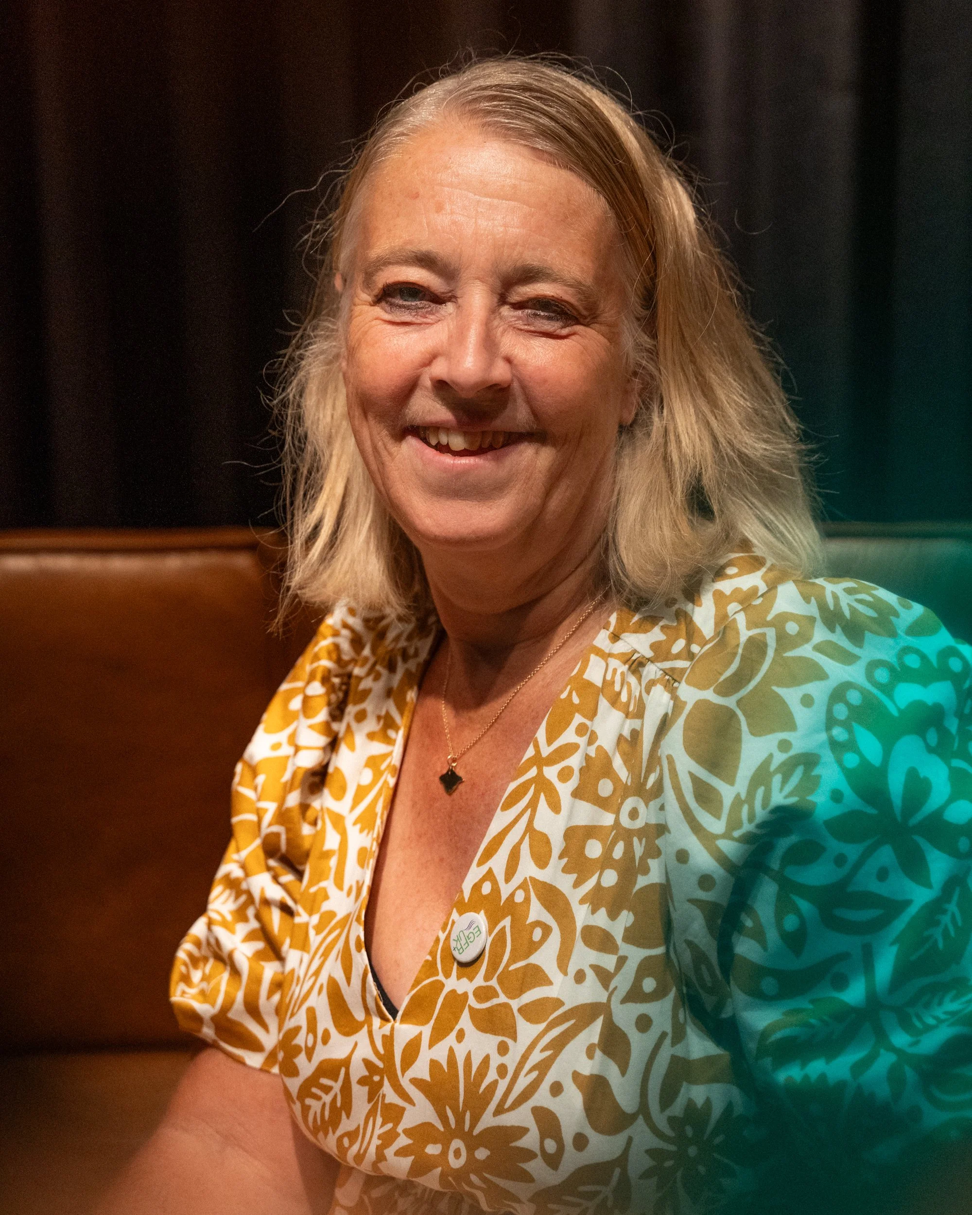 Smiling woman with blonde hair wearing a patterned shirt and a button that says "EACH."