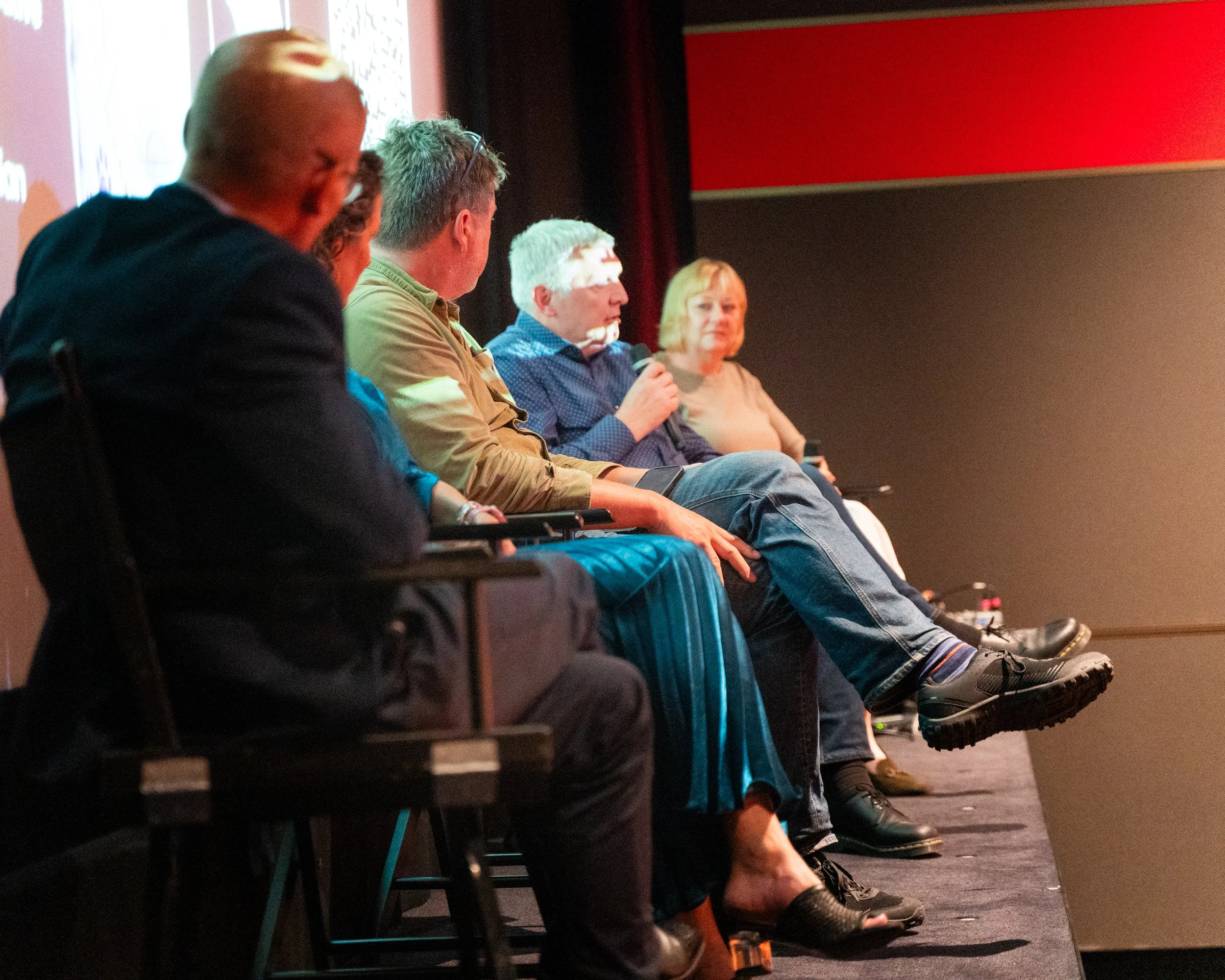 People seated in a row on chairs facing a stage, with some wearing glasses, holding microphones, and looking in the same direction. The background shows a projection screen and a red wall.