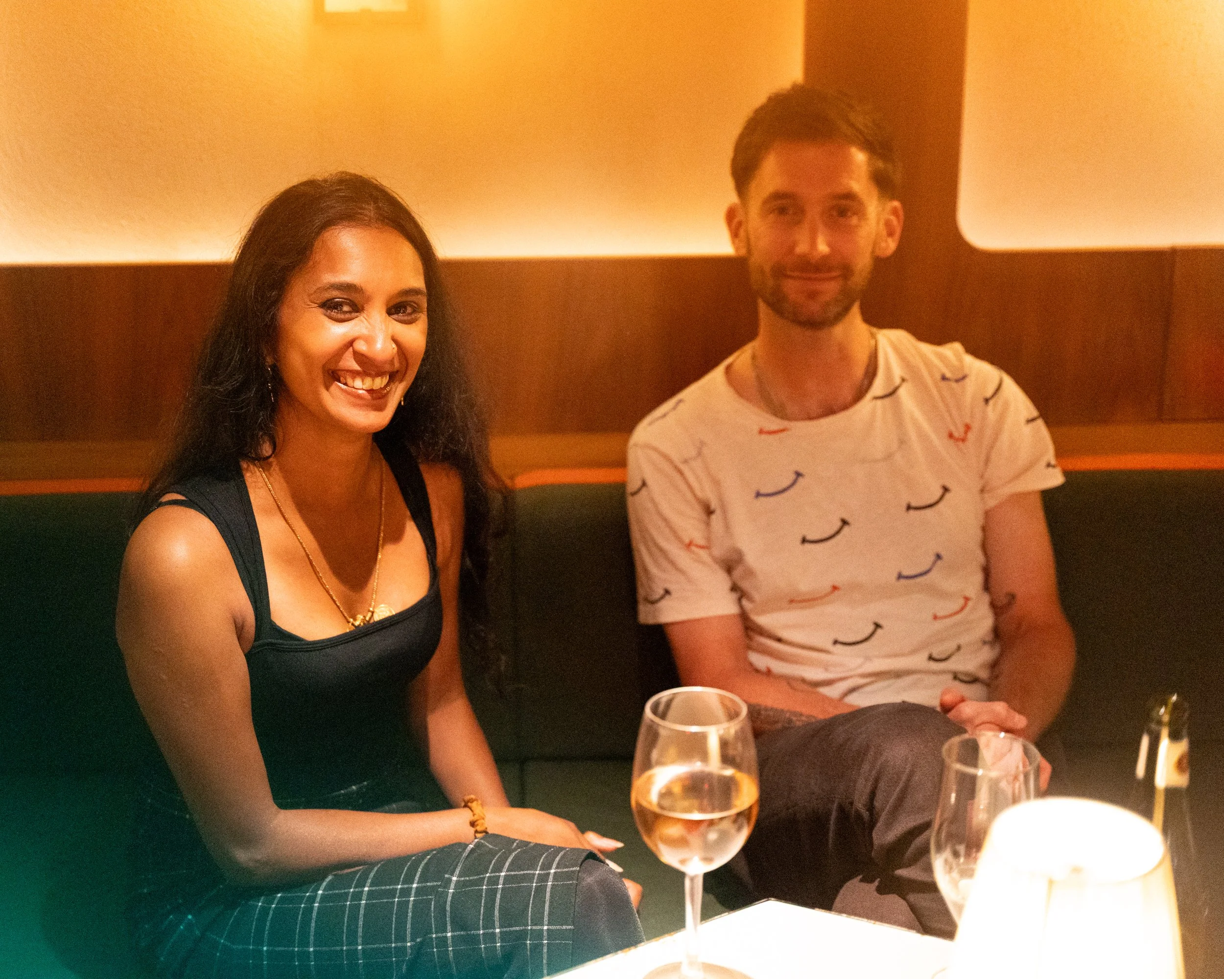 A smiling woman with dark hair and a nose piercing sitting at a table with a man in a white shirt with a mustache pattern. There are wine glasses on the table in front of them, in a warmly lit restaurant.
