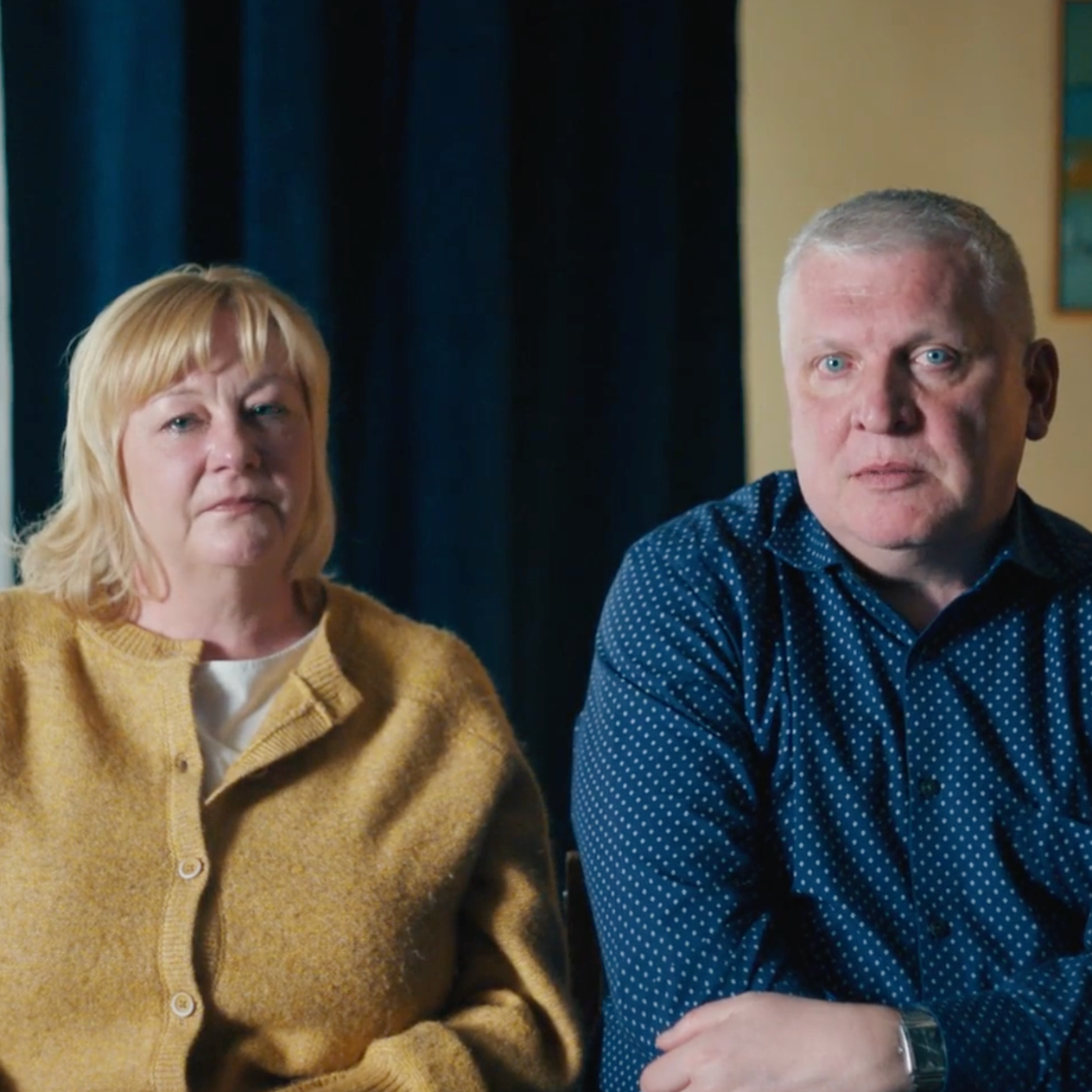 A middle-aged woman with blonde hair wearing a mustard-colored sweater and a middle-aged man with short blonde hair wearing a blue patterned shirt, both looking serious, seated indoors with dark curtains and a yellow wall in the background.