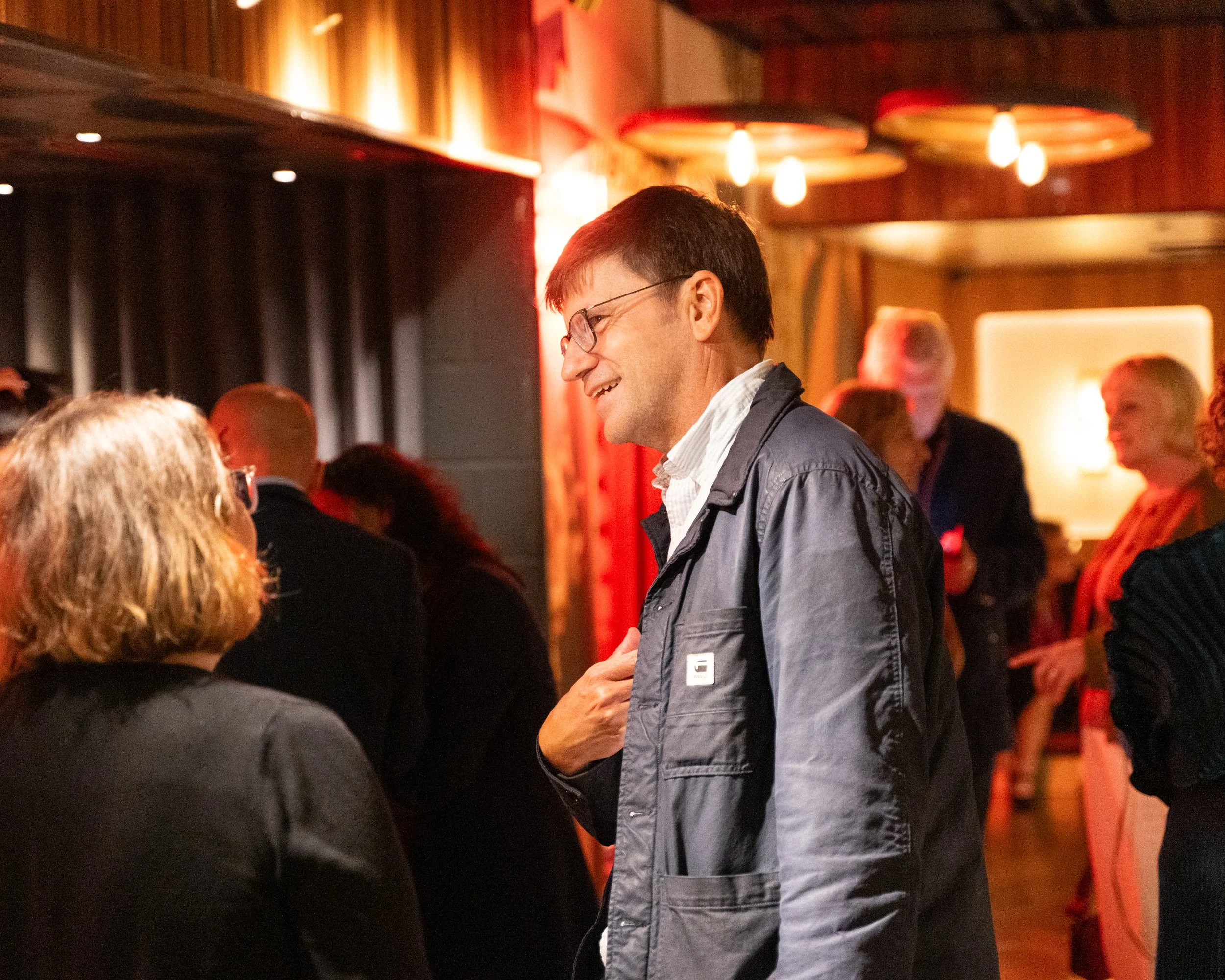 A man wearing glasses and a gray jacket smiling and talking to a woman with blonde hair in a crowded indoor setting with warm lighting and wooden decor.