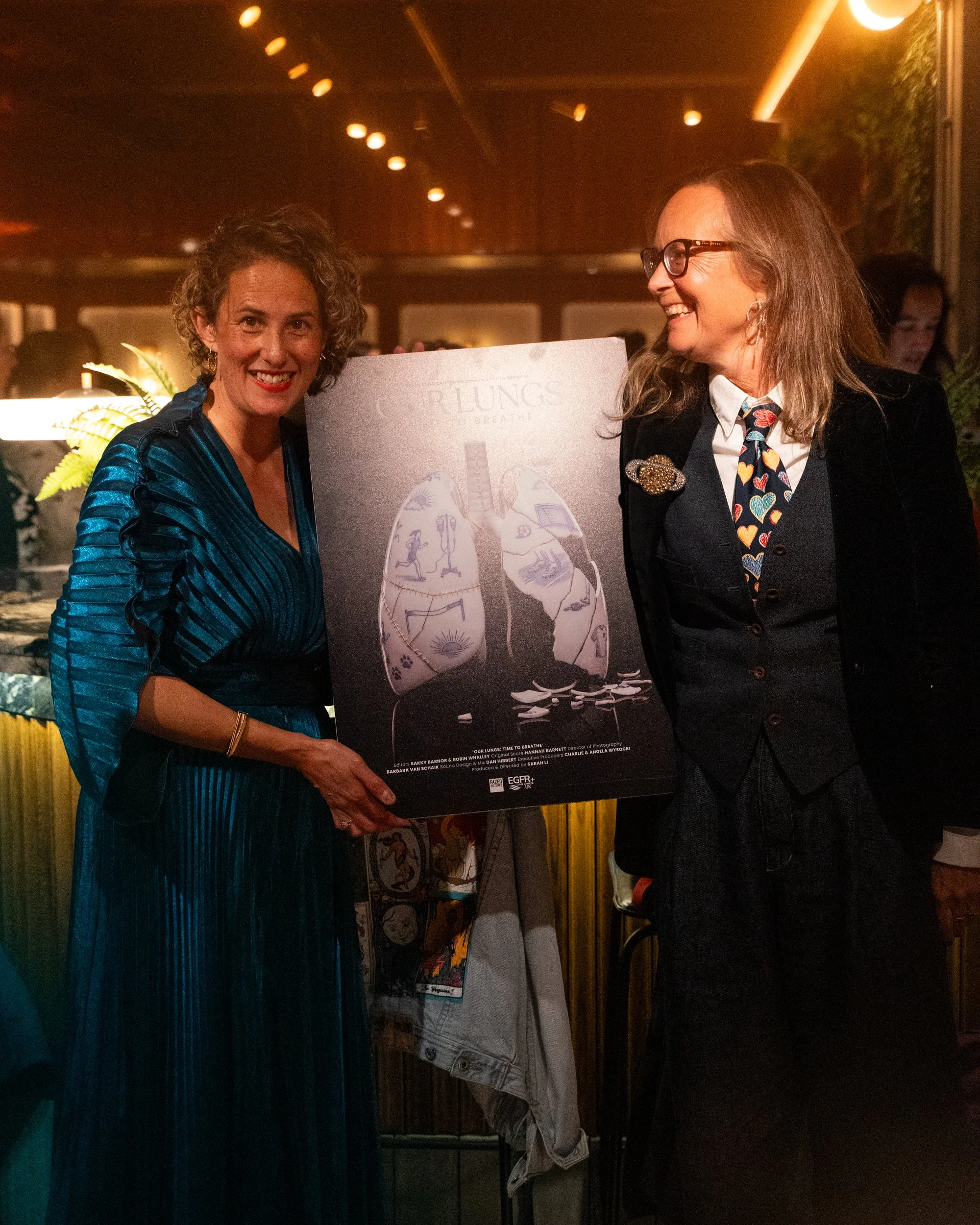 Two women smiling and holding a poster of human lungs at an indoor event, with warm lighting and other people in the background.