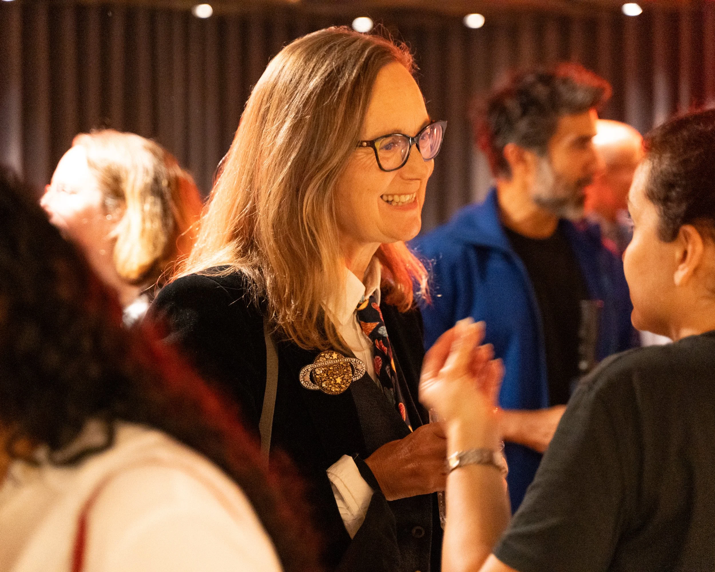 A group of diverse people talking and laughing at an indoor social gathering with warm lighting.