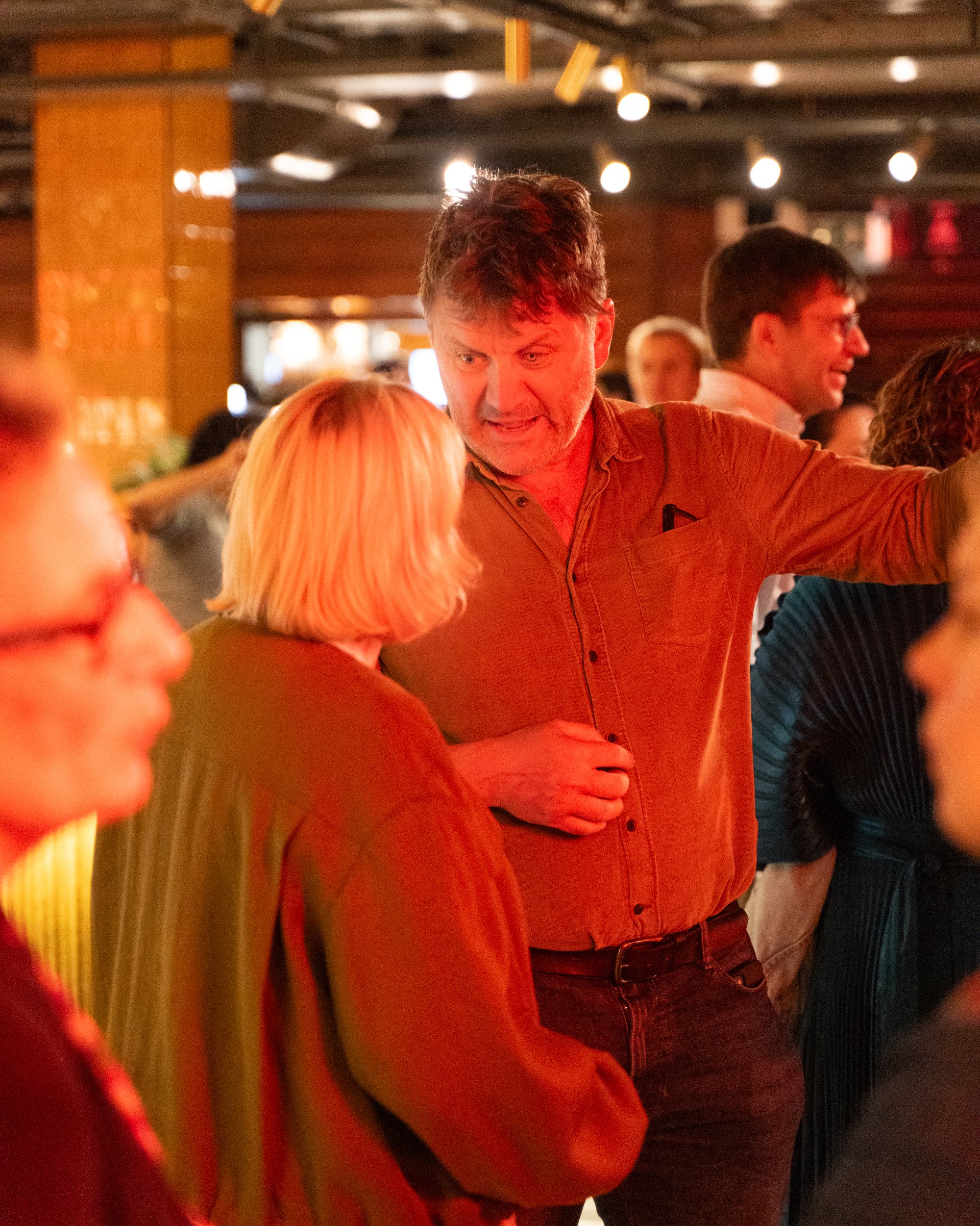 People socializing at a dimly lit indoor venue, with a man with curly dark hair and light beard in brown shirt speaking to a woman with blonde hair, other individuals in the background.