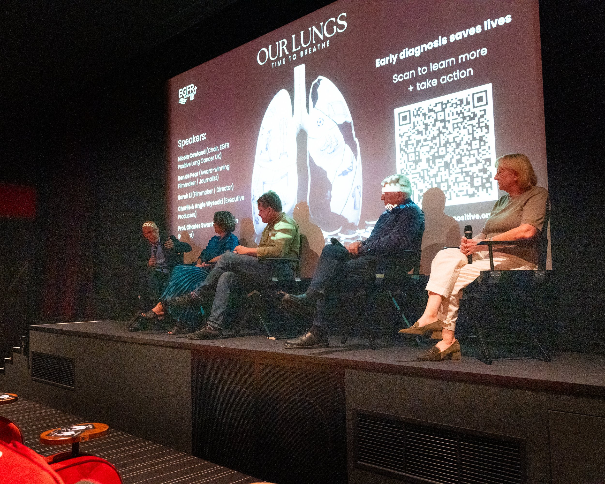 Panel of five people seated on stage at a panel discussion during a conference, with a large screen behind them displaying the event's branding and a QR code for more information.
