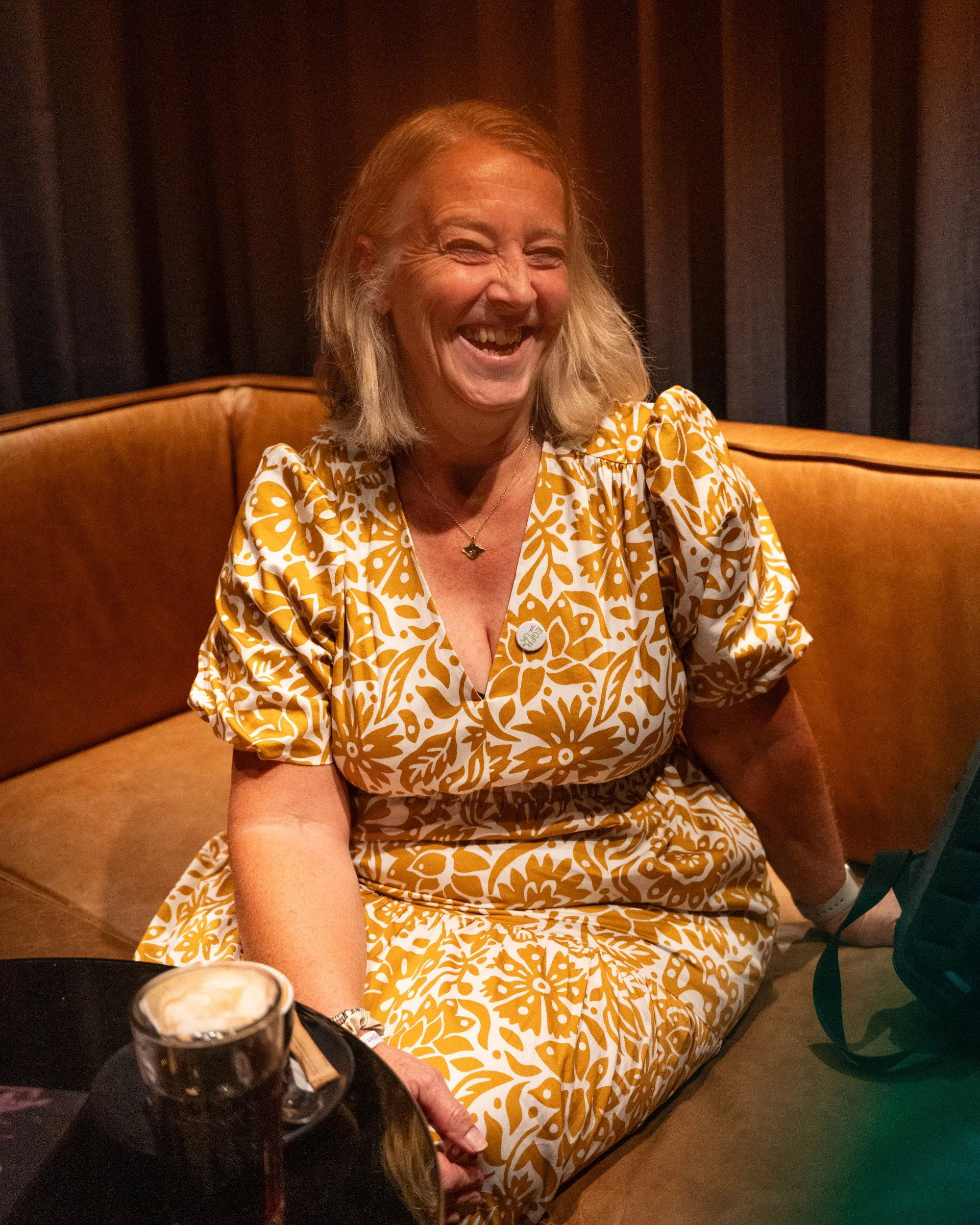 A woman with blonde hair, wearing a yellow and white patterned dress, is sitting in a booth and laughing. There is a bottle of beer on the table in front of her and a partially visible drink with whipped cream. The background has dark curtains.