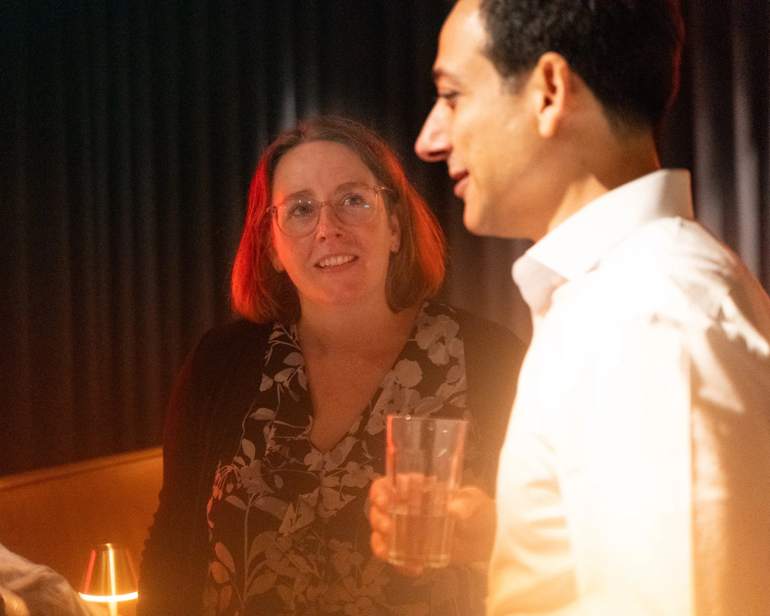 A woman with glasses and red hair looking at a young man with short dark hair, both engaging in a conversation in a dimly lit setting, possibly a bar or restaurant, with a drink in front of them.