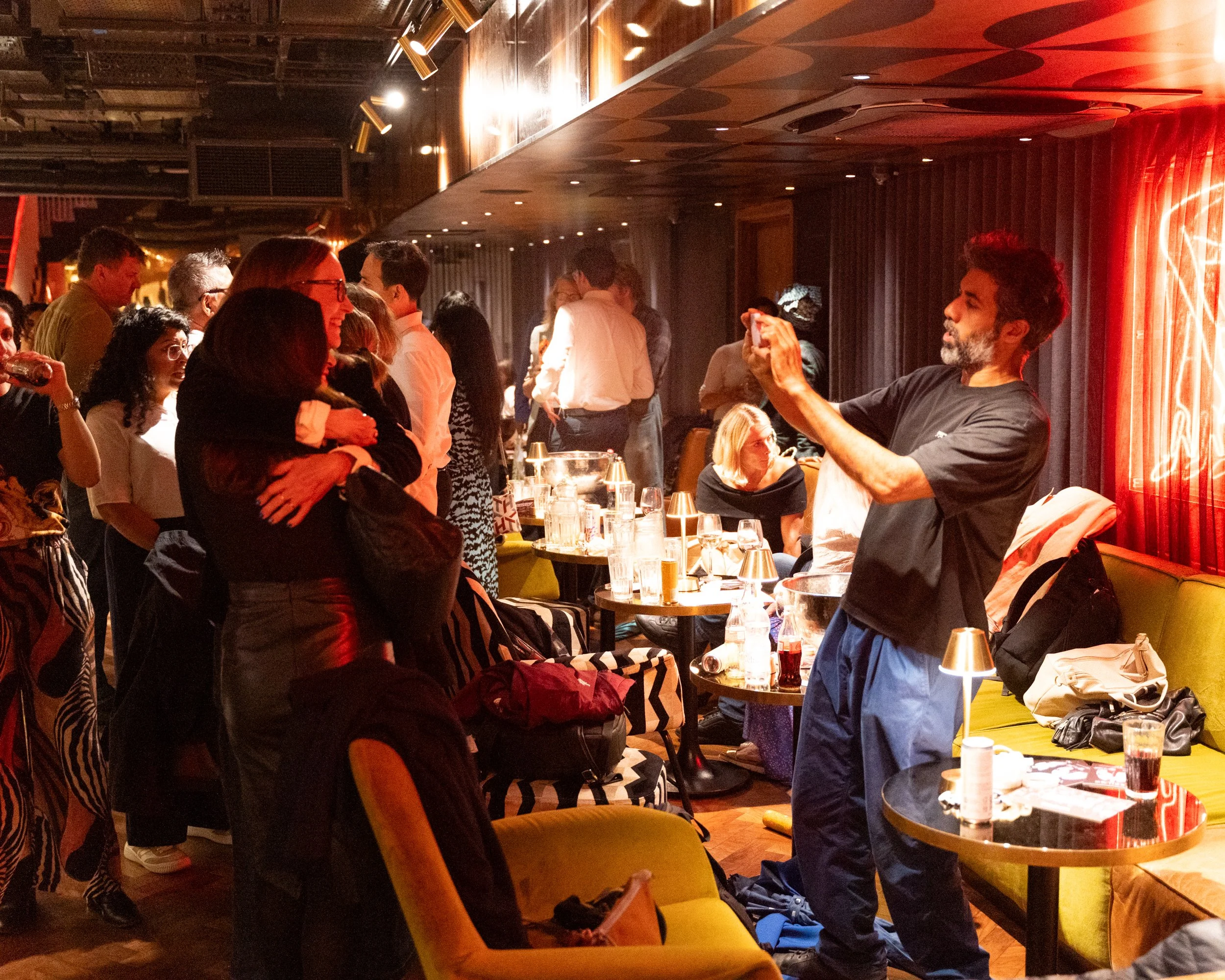 People gather at a social event in a dimly lit room with ambient lighting, some standing and engaging in conversations while others are seated at tables with drinks and food, and a man is taking a photo of the group.