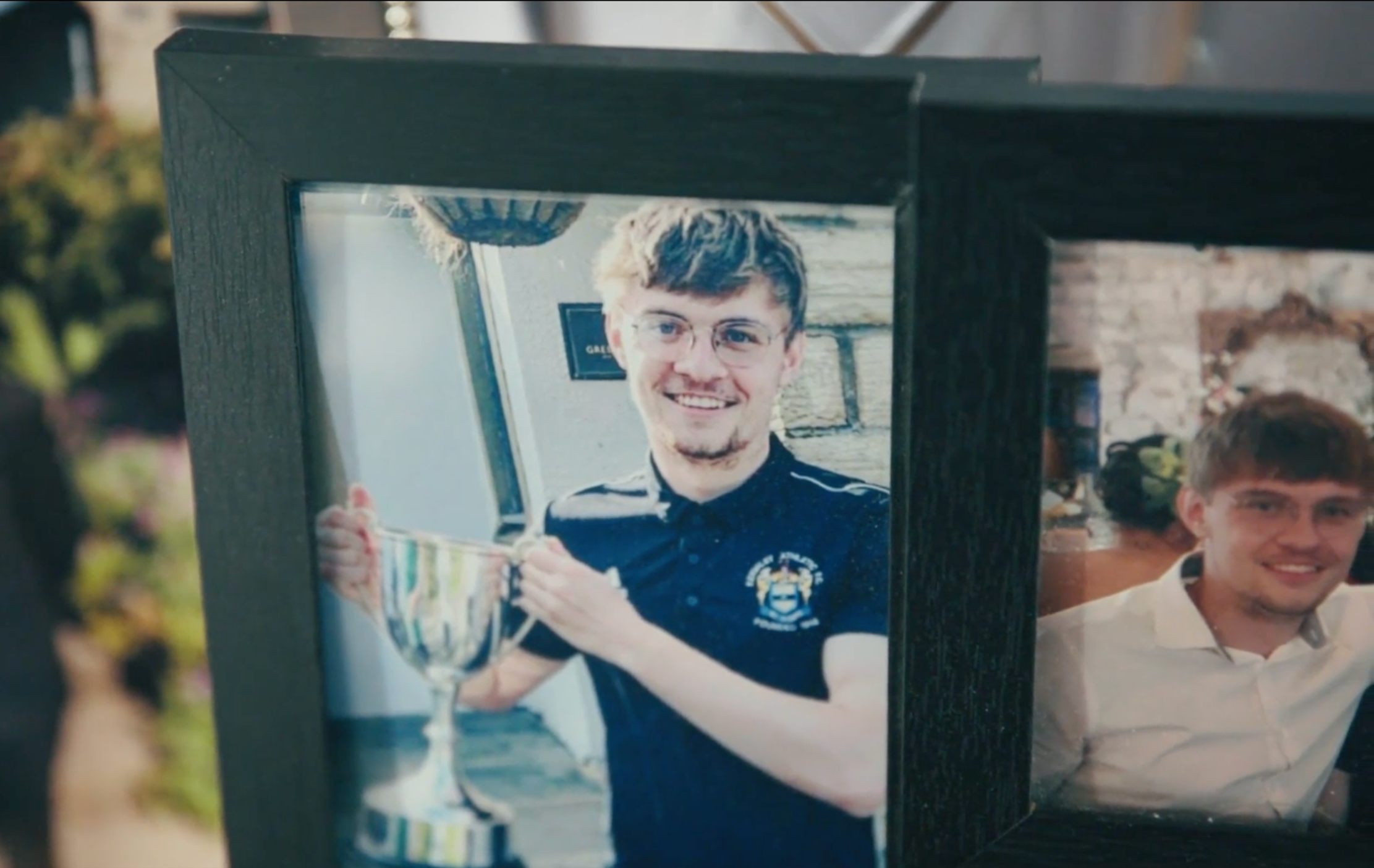 Alex Wysocki, a young man with glasses holding a trophy and smiling, seen through a framed photograph on a black wooden surface inside a building. Alex died from EGFR+ lung cancer aged 24.