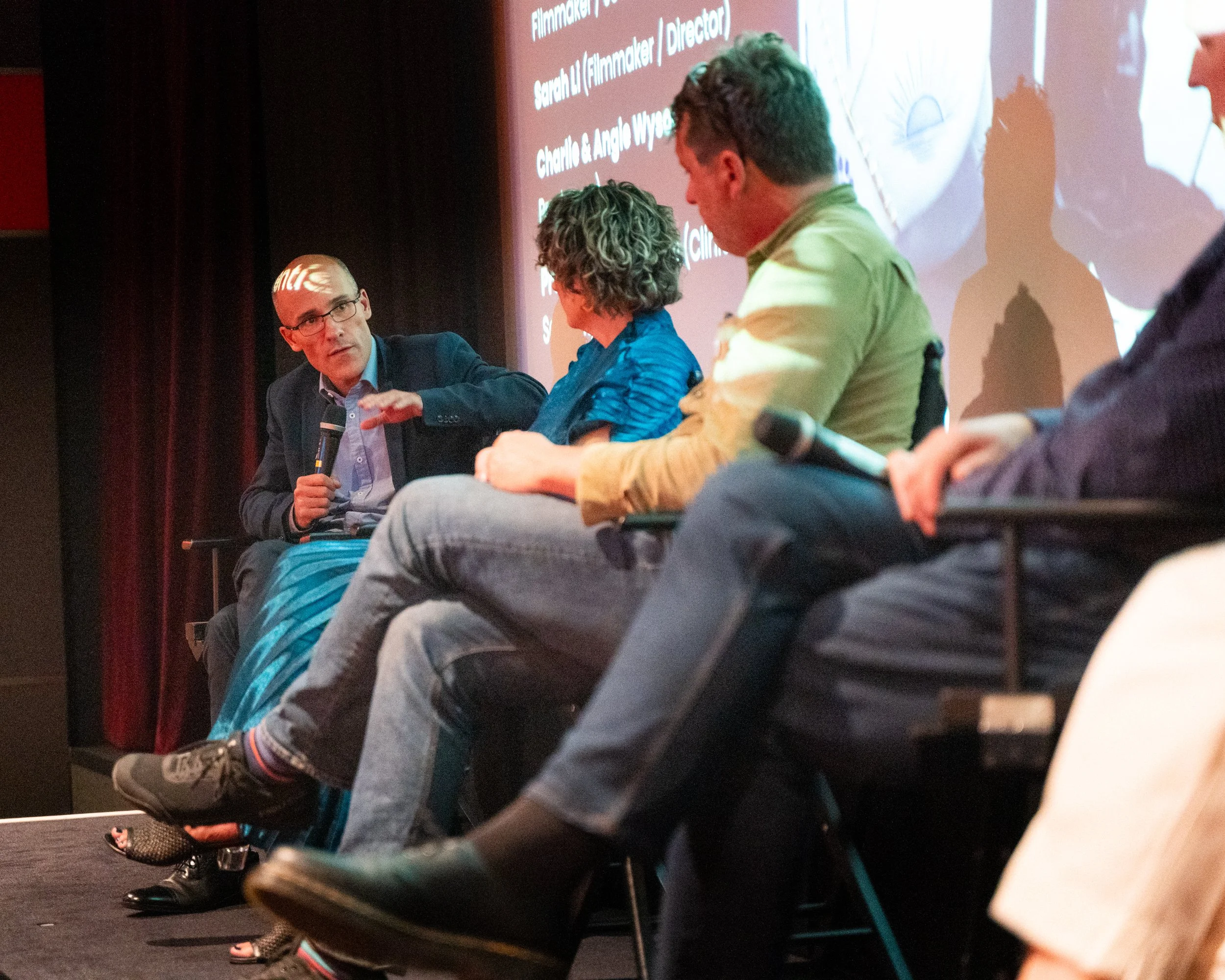 The image shows a panel discussion with five individuals seated at a table. One man is speaking into a microphone, wearing glasses and a dark blazer. The others are listening attentively. A large screen behind them displays text related to a film eve