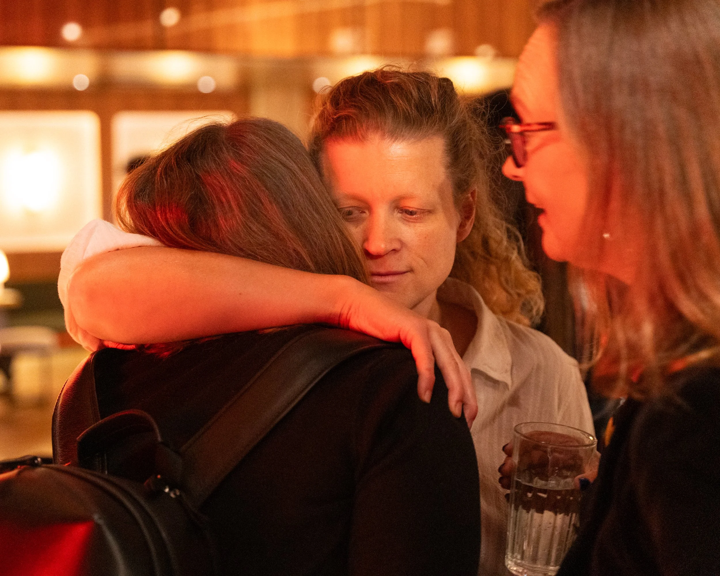 Three women sharing an affectionate hug at a warmly lit indoor gathering, with one holding a glass of wine and another holding a glass of water.