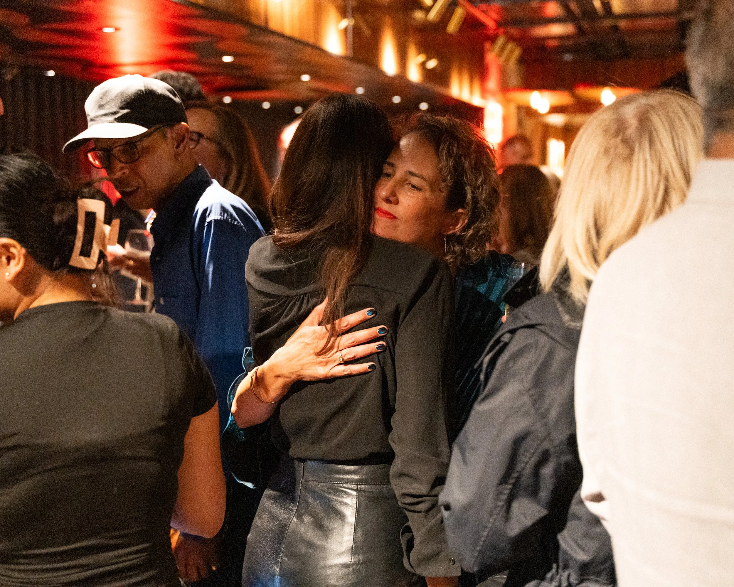 People socializing at a dimly lit indoor gathering or party, embracing and chatting.