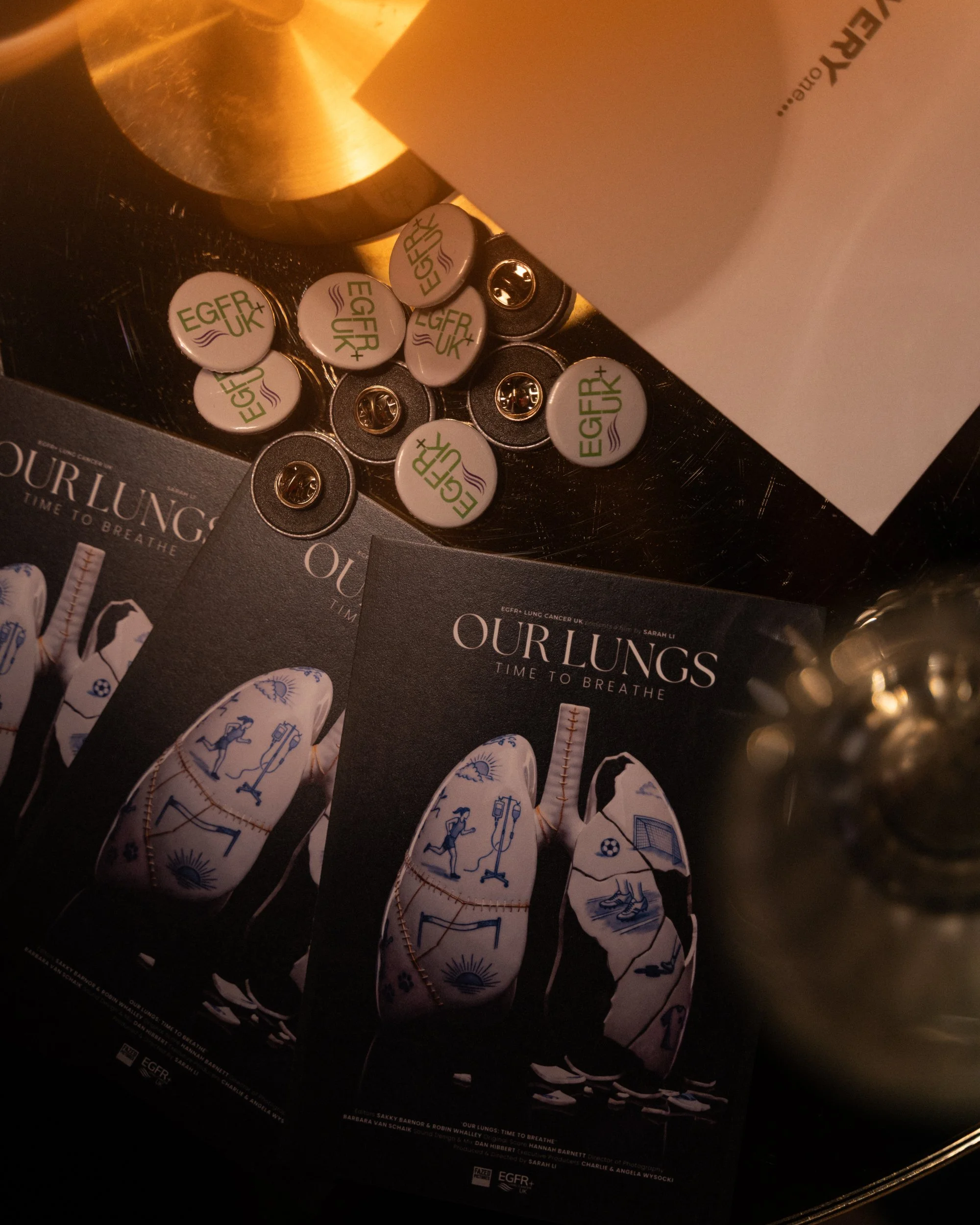 Close-up of a black magazine titled 'Our Lungs: Time to Breathe,' with scattered white circular stickers bearing the logo 'EGFR+ UK' in blue and green. Part of a golden-colored object is visible in the upper left corner, and a white paper with partia