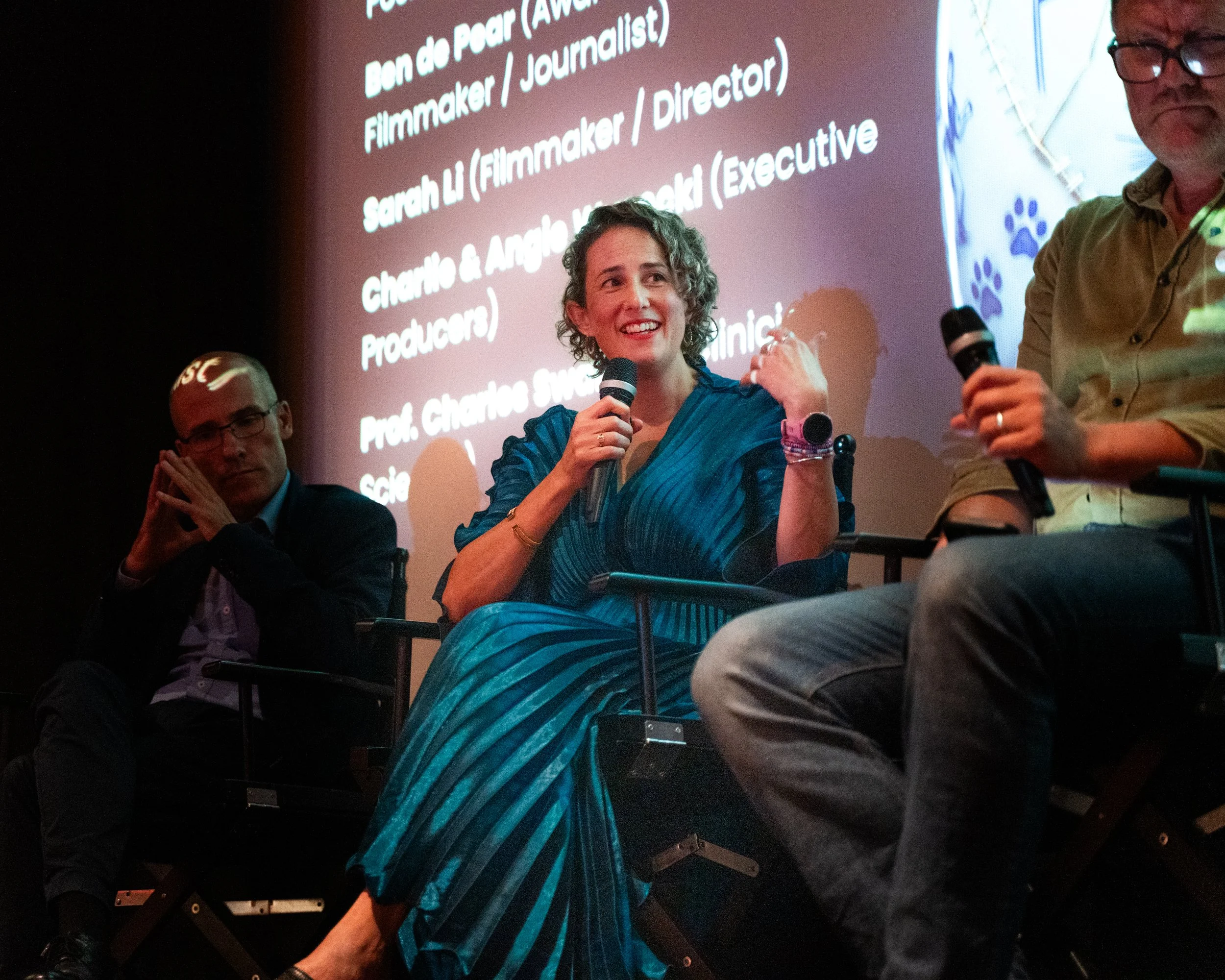 A woman with curly hair and a blue dress speaking into a microphone at an event. Behind her, a large screen shows a list of names and titles including 'Ben de Poor,' 'Sarah Li,' and 'Charlotte Swenson.' Two other men are partially visible, one holdin